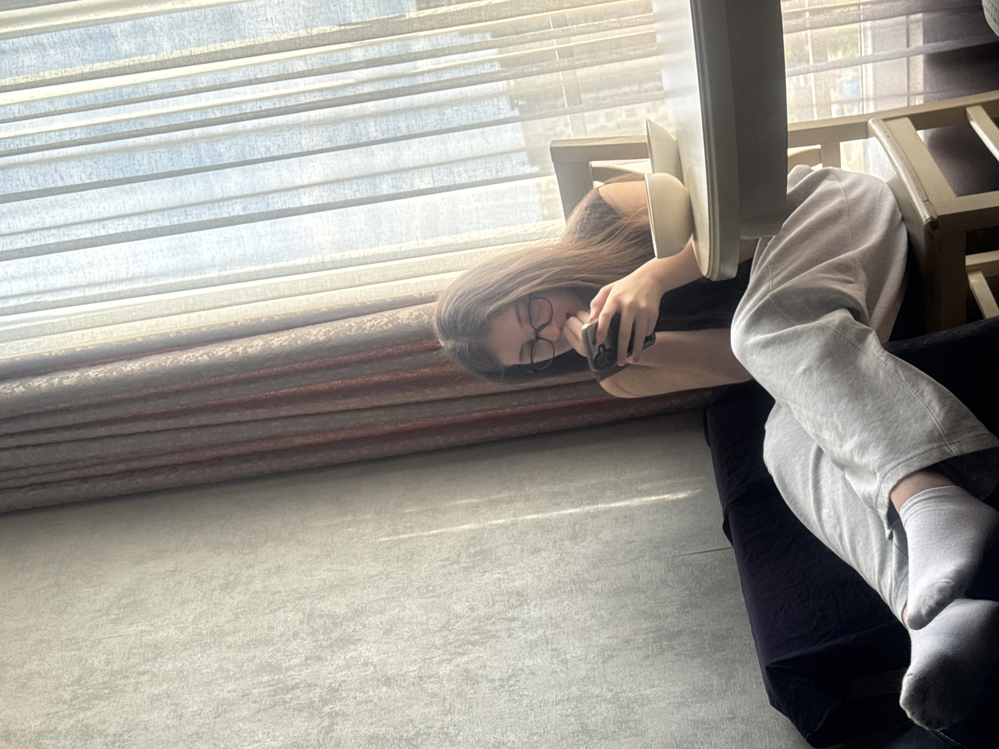 A young woman with long brown hair and glasses is reclining on a dark-colored sofa in what appears to be a room with a window covered by blinds. She is holding a smartphone in her hand and looking at it. The woman is wearing gray sweatpants and white socks. A wooden chair is partially visible near the sofa. The environment is softly lit, likely by daylight filtering through the blinds. The location is presumed to be in a residential area within the city of Tbilisi, Georgia, based on available information.