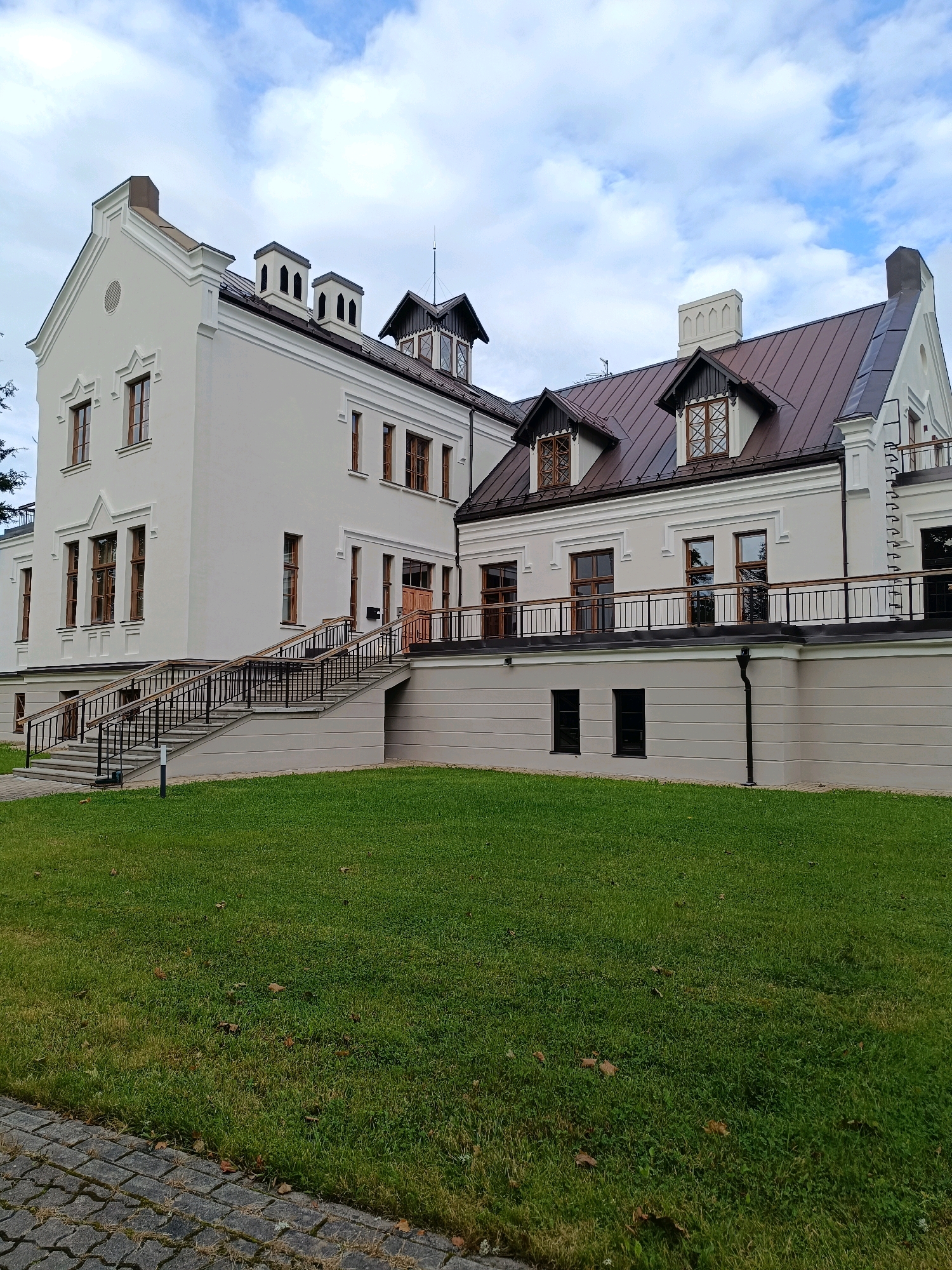The image shows a large, modern, two-story building with a light-colored facade and a dark brown metal roof. The building has multiple windows, some with decorative cross-hatching in the upper panes, and several chimneys. A wide set of stairs with black railings leads up to the main entrance on the left side of the building. A black metal railing runs along a raised patio area that encircles a portion of the building. The ground in front of the building is covered by a lush green lawn with scattered brown leaves. To the left of the stairs, a cobblestone walkway is visible. The sky is partly cloudy with patches of blue. The building appears to be located in Bojas, Latvia.