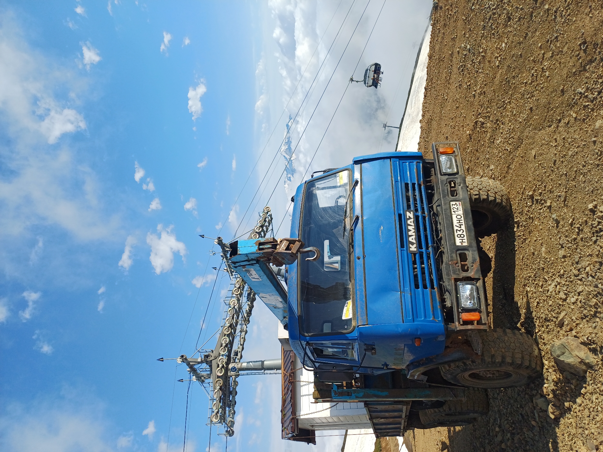 A bright blue KAMAZ truck with a crane attachment is parked on a gravelly, mountainous terrain. The truck is facing forward and appears to be a utility vehicle. In the background, a ski lift structure with cables and chairs is visible, with some chairs occupied by people. Snow patches are present on the ground, suggesting a high-altitude location. The sky is blue with scattered white clouds, and distant snow-capped mountains are visible above a layer of clouds. The scene is likely in or near Estosadok, Russia, a mountain resort town known for its ski lifts and mountainous landscape.