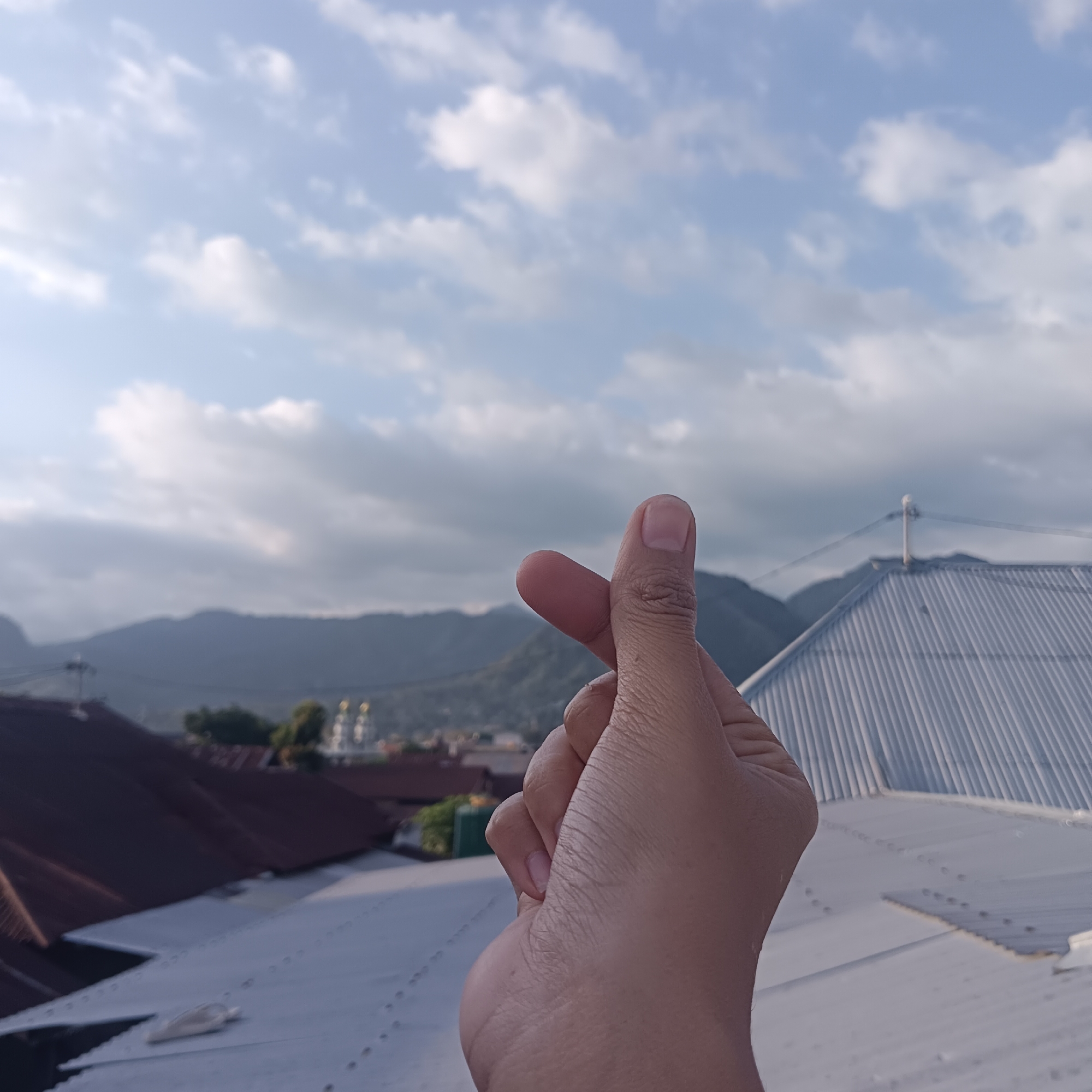 The image shows a close-up of a person's hand making a Korean heart gesture against a blurred background of rooftops, mountains, and a cloudy sky. The hand is positioned in the foreground, with the thumb and index finger touching to form a heart shape. The rooftops are varied in color and material, with some appearing to be made of corrugated metal. In the distance, rolling hills and mountains are visible, shrouded in a light haze. The sky is a pale blue with scattered white clouds. The scene suggests an outdoor setting, possibly on a rooftop overlooking a town or city. The location is identified as Paupanda, Indonesia.