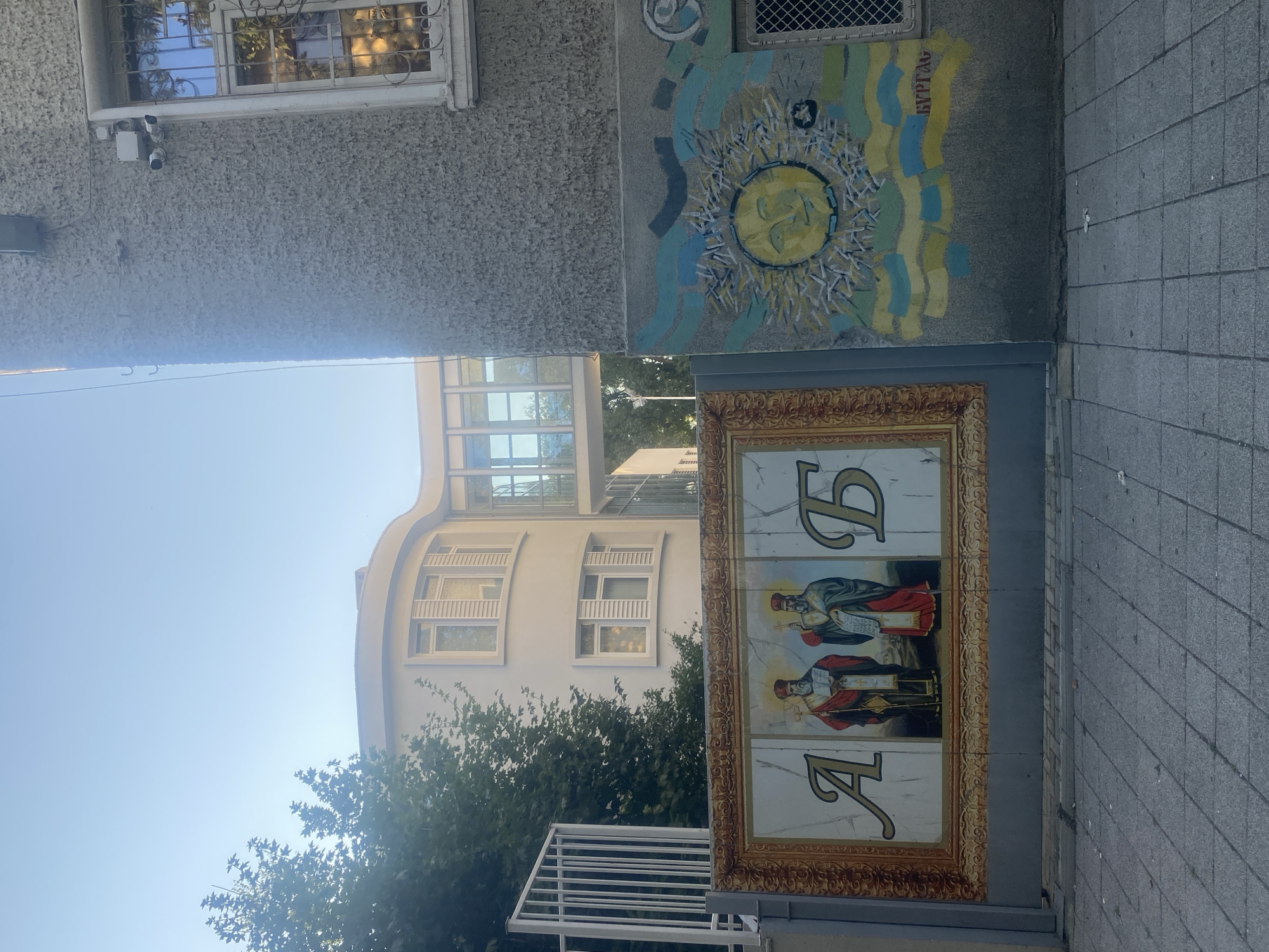 The image shows a street scene in Burgas, Bulgaria. On the left, there is a large gate or mural painted to resemble a gilded frame. Inside the frame are two religious figures in traditional attire, holding staffs and books. Large, ornate Cyrillic letters are visible on either side of the figures. To the right of the gate, the wall of a building is covered in a colorful mural depicting a smiling sun with rays and wavy blue and yellow patterns, reminiscent of the Ukrainian flag. The word "БУРГАС" (Burgas) is written in yellow on this mural. Behind the gate and to the left, a modern, light-colored building with curved architectural elements and multiple windows is visible, partially obscured by trees. To the right, a rough-textured concrete wall of another building has a window with decorative iron bars. Security cameras are mounted on this wall. The ground in the foreground is paved with gray square tiles. The sky is clear and blue.