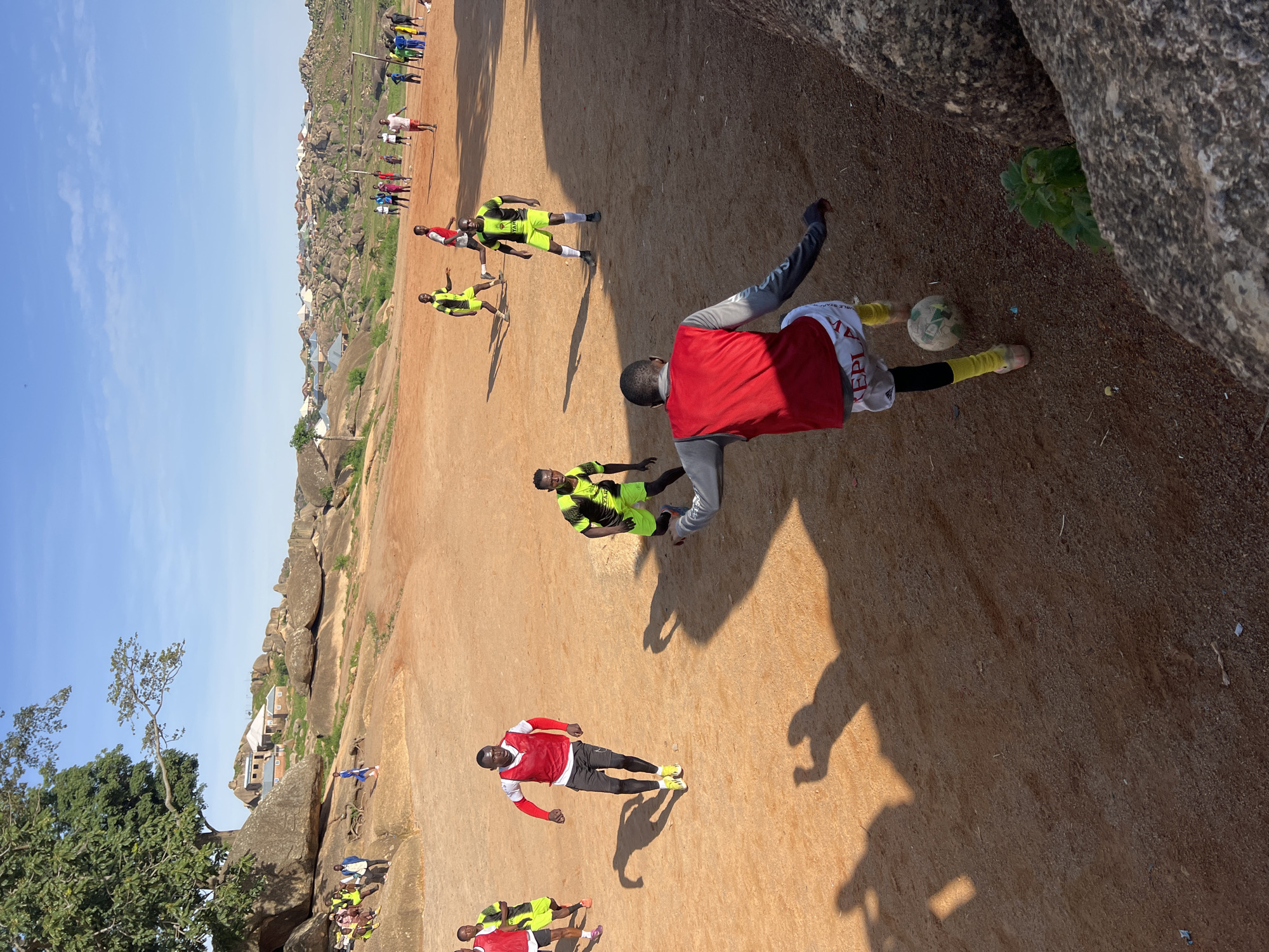 The image depicts an outdoor soccer game being played on a dirt field in the city of Jos, Nigeria. The field is characterized by its reddish-brown dirt surface, with large rock formations scattered throughout the landscape and along the edges. In the background, a settlement with houses built among the rocky hills is visible, indicative of the rugged terrain of Jos.
Several individuals are actively engaged in the soccer match. Most players are wearing athletic attire, including shorts and jerseys or bibs. Some are in motion, kicking or running with the soccer ball, while others appear to be positioned on the field, observing the game or anticipating play. A soccer ball is visible on the ground, being kicked by one of the players in the foreground. The players' shadows are cast long on the dirt field, suggesting it is either early morning or late afternoon.
The environment is sunny with a clear blue sky. The presence of large rocks and the style of the buildings in the background are consistent with the geological and architectural features commonly found in Jos. The overall scene conveys a sense of local community activity and sport in a natural, somewhat arid setting.