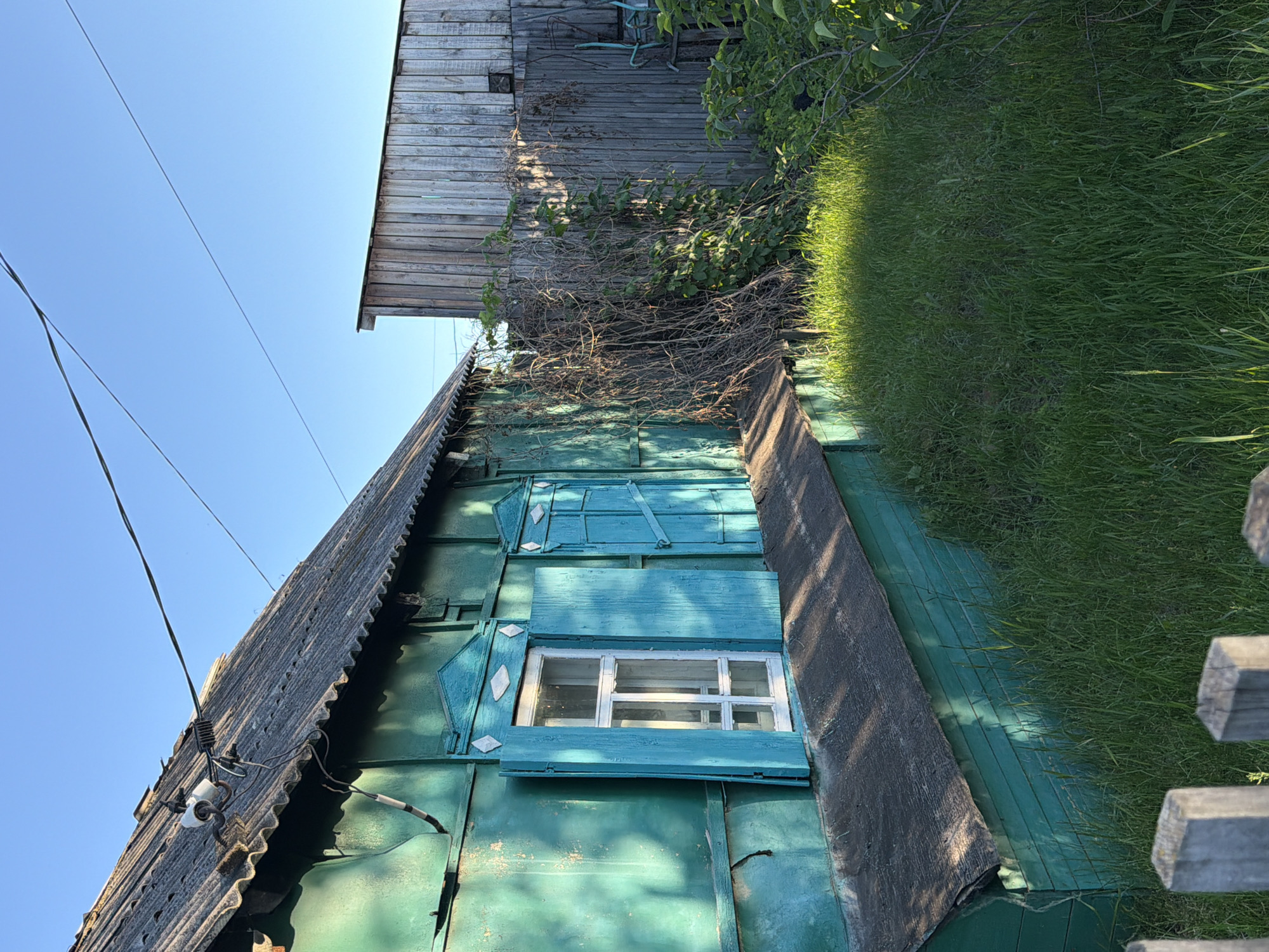A weathered, teal-painted wooden house with a gray corrugated roof is visible under a clear blue sky. The house features a white-paned window with open, teal shutters. A dark, sloped foundation runs along the base of the house, which is surrounded by lush green grass. To the right of the house, a tall, weathered wooden fence is partially obscured by dense, dry branches and green foliage. Overhead, power lines stretch across the sky. The scene appears to be a residential property in the city of Kansk, Russia, likely in a rural or semi-rural area, given the unkempt natural elements and the traditional style of the house and fence. There are no people or animals present in the image.