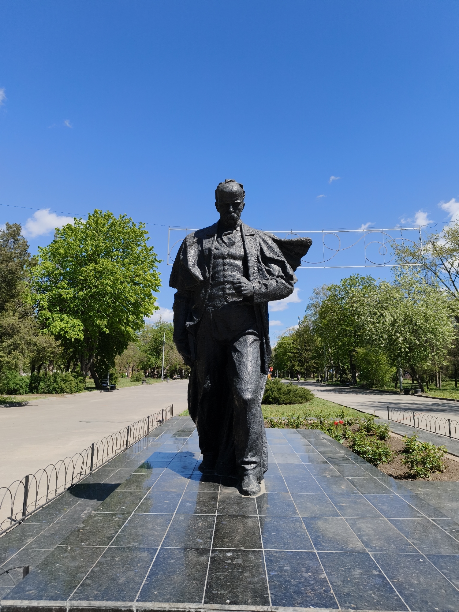The image captures a full-length bronze or dark metal statue of a man standing prominently in what appears to be a park or public square in Kryvyi Rih, Ukraine. The man in the statue is depicted with a beard and mustache, wearing a suit with a long coat or cloak draped over his left shoulder. He stands on a polished black tiled pedestal, which reflects the clear blue sky and surrounding greenery. A low, decorative metal fence encircles the base of the monument, separating it from a paved pathway and a small flowerbed containing green shrubs and a few red tulips.
Behind the statue, a wide paved avenue stretches into the distance, flanked by numerous trees with vibrant green foliage, suggesting springtime. The trees vary in density and shade of green, indicating different species. In the mid-ground, a decorative metal structure with curved patterns is suspended across the avenue, possibly for banners or lights. The scene is brightly lit by natural sunlight, casting distinct shadows on the tiled base and along the path. The sky above is a brilliant, clear blue with a few scattered white clouds, indicating sunny weather. The overall setting is serene and verdant, typical of an urban park on a pleasant day. No people are visible other than the statue, and no text is discernible in the image.