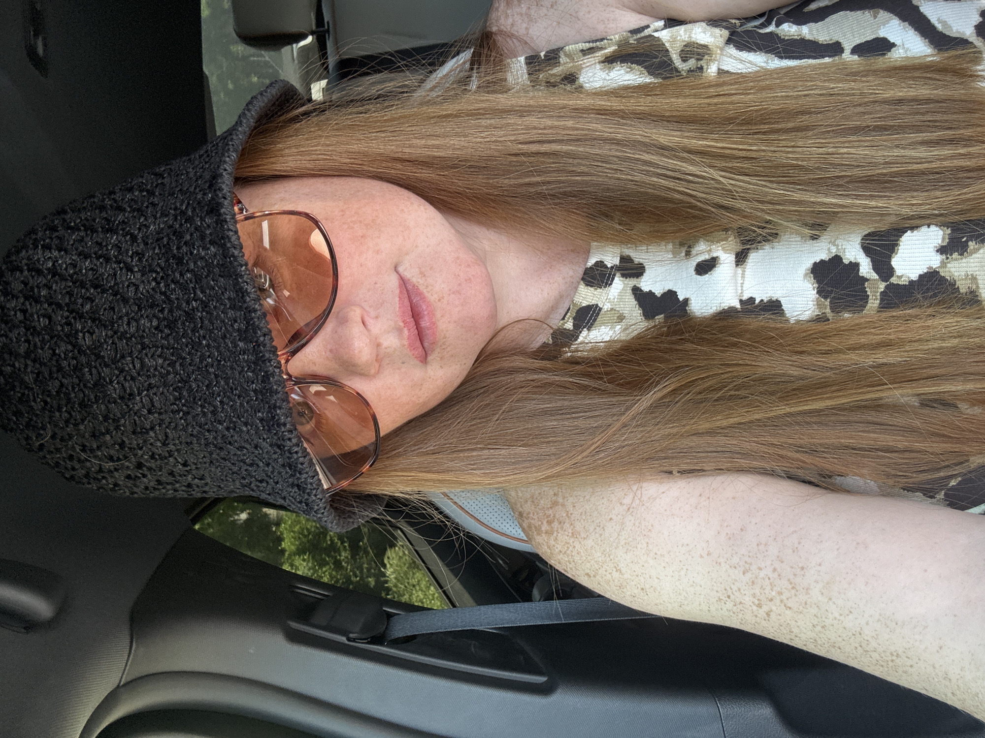 A woman with long red hair and freckles is sitting in the passenger seat of a car. She is wearing a black crocheted bucket hat and rose-tinted sunglasses, with her head tilted forward. She is also wearing a sleeveless top with a leopard-print pattern. The interior of the car is visible, including a seatbelt and part of the car's headrest. Through the car window, trees and foliage are visible, suggesting an outdoor location. The image appears to be a selfie taken by the woman. There is no indication of the city of Yerevan, Armenia.