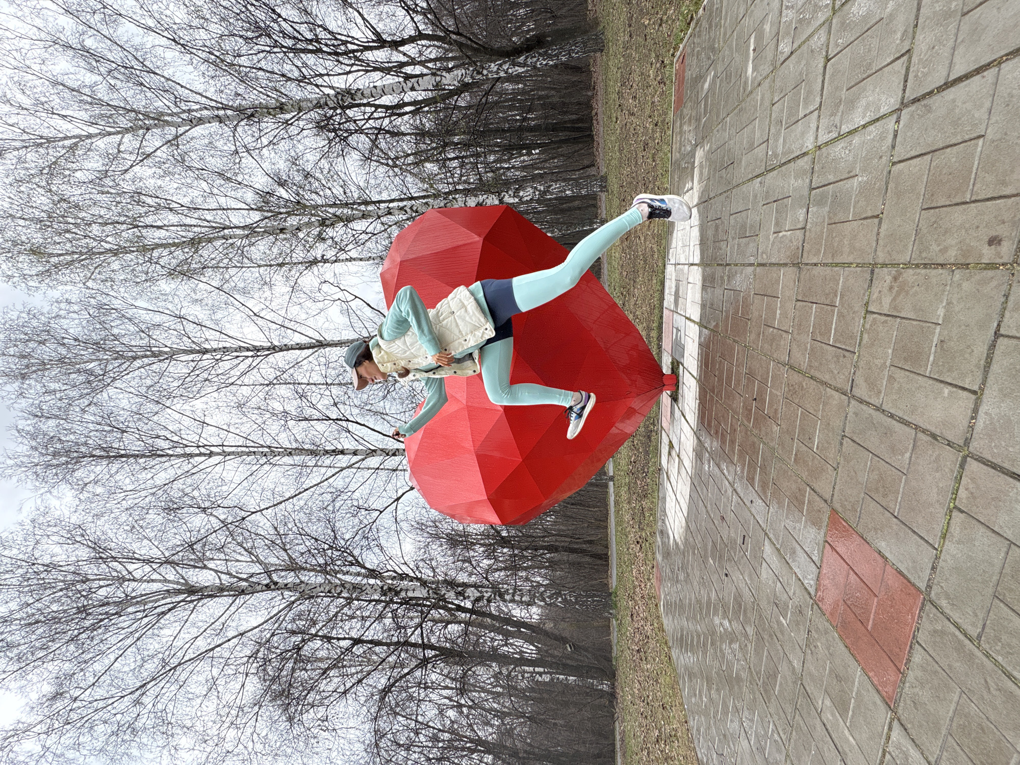A woman stands on a brick pavement next to a large, red heart-shaped sculpture. She is wearing a light-colored jacket, blue leggings, and athletic shoes. Her left leg is extended upward. The background shows bare trees, indicative of late autumn or early spring. The pavement is wet, possibly from recent rain. This scene takes place in Ulyanovsk, Russia.