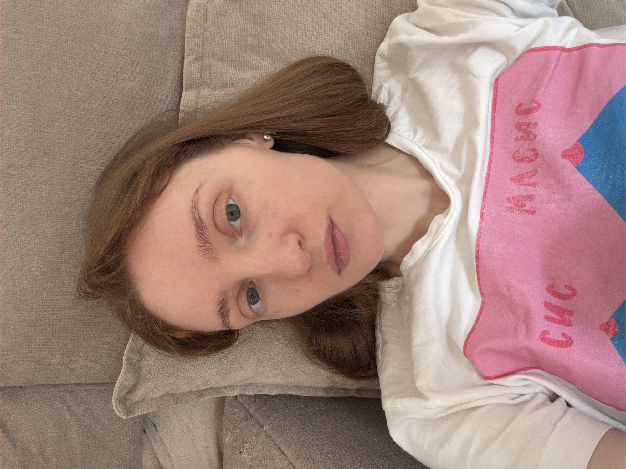 A young fair-skinned woman with auburn hair is lying on a beige upholstered sofa or cushion. She is wearing a white t-shirt with a pink and blue graphic design. The image is taken from a high angle, looking down at her. Her head rests on the cushion. The background is out of focus, but appears to be the sofa's surface. No other people are visible. Location details are not discernible from the image. The description omits mention of Boreti, Montenegro, as there is no information to suggest this location.