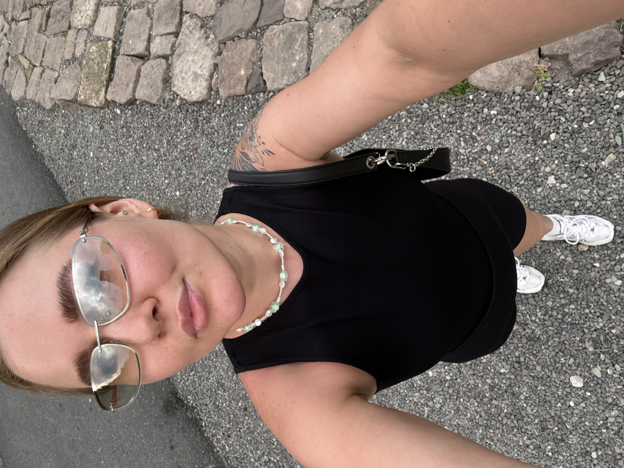A woman takes a selfie in an outdoor environment. The location appears to be a walkway or path, with stone paving along one side and a gray, gravel-like surface making up the majority of the ground. She is wearing a black dress, a beaded necklace, and white sneakers. She has glasses on and is wearing a black belt-bag. There is a tattoo visible on her left arm. Based on the environmental details and lack of specific landmarks, definitive identification of the exact spot in Jinhua, China, is not possible.
