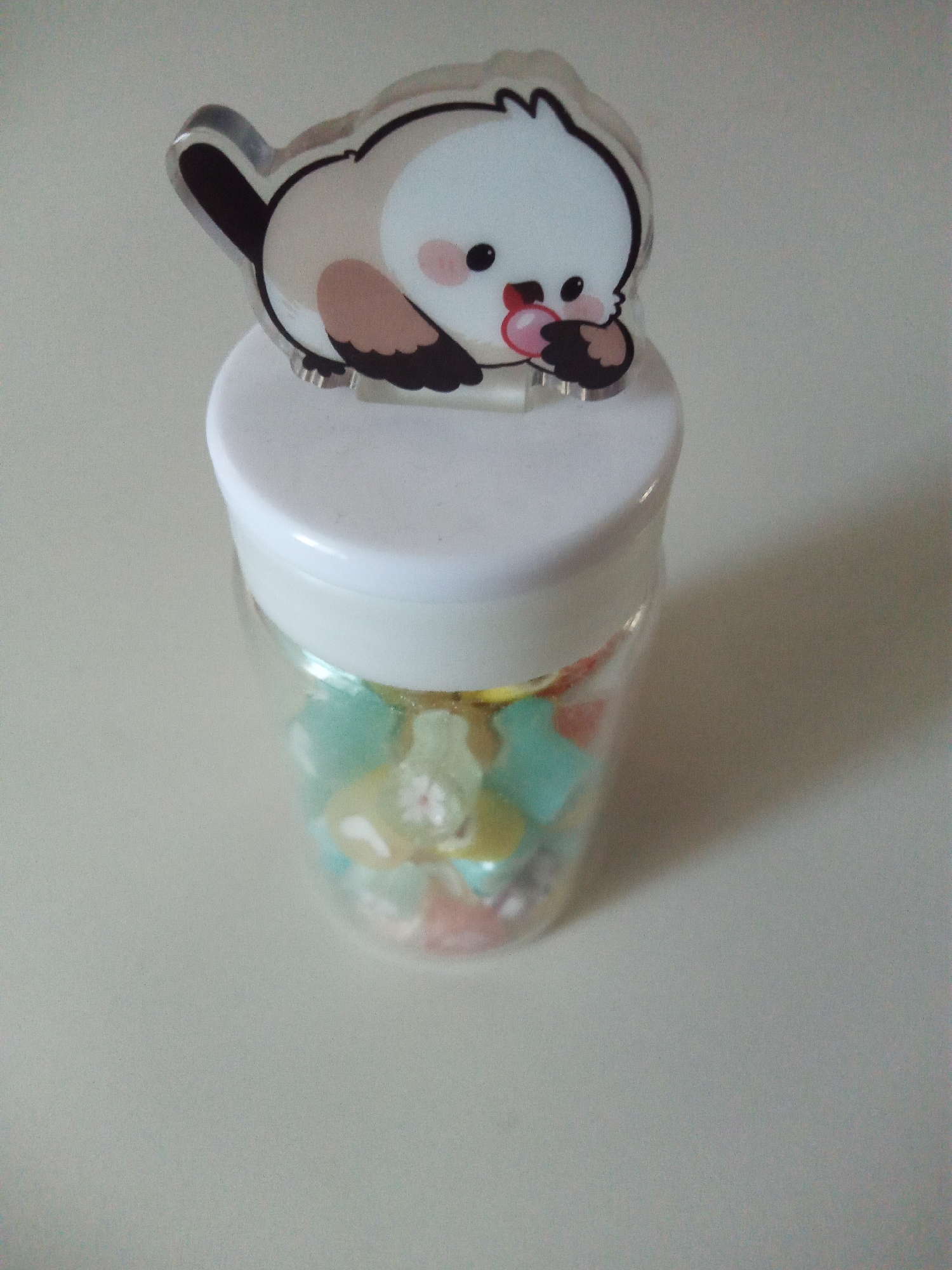 The image shows a small, clear glass jar with a white lid. The jar is filled with colorful, translucent candies in various shapes, including stars and possibly other small figures. A decorative acrylic cutout of a cute, cartoonish bird is perched on top of the jar's lid. The bird is depicted with a white and beige body, brown wings, and a pink cheek. It is holding a pink heart-shaped object in its beak. The background is a plain, light-colored surface, possibly a table, with subtle shadows indicating a light source. The overall scene is simple and appears to be a close-up shot of a decorative item. There is no visible text in the image. The time of day and weather are not discernible from the image. The location context of Usman, Russia is provided but has no direct visual cues within the image itself.