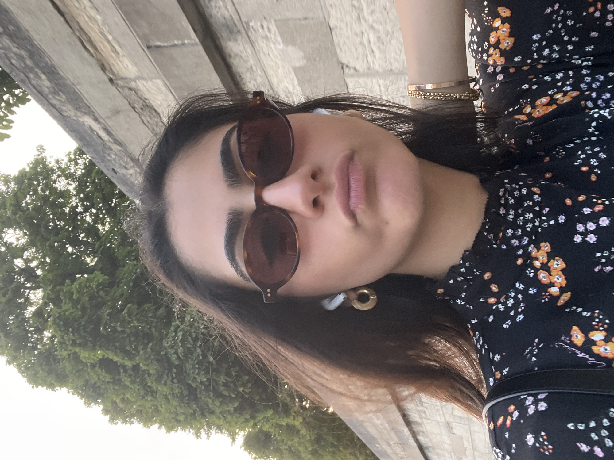 A young woman with dark hair and sunglasses is taking a selfie outdoors. She is wearing a black dress with a floral pattern, gold hoop earrings, and AirPods. She has on a bracelet on her right arm. The background shows a stone wall and trees, with a hint of a pale sky. The lighting suggests it is either early morning or late afternoon. There is no specific indication of the city of Paris, France within the image.