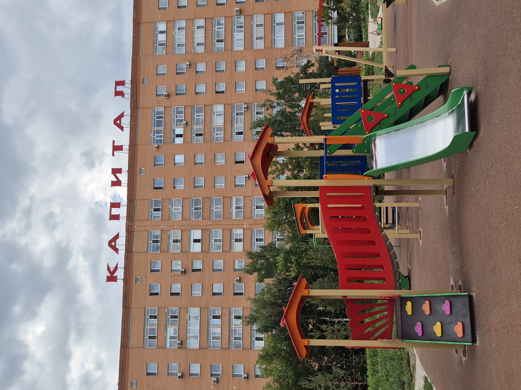 An outdoor view of a brick apartment building with red lettering spelling "КАПИТАЛ" across the top. The building has many windows, some with air conditioning units mounted below them. In front of the building is a children's playground with colorful slides, climbing structures, and swings. The ground of the playground is a reddish-brown rubber surface. Trees and greenery are visible behind the playground. The sky is overcast with gray clouds.