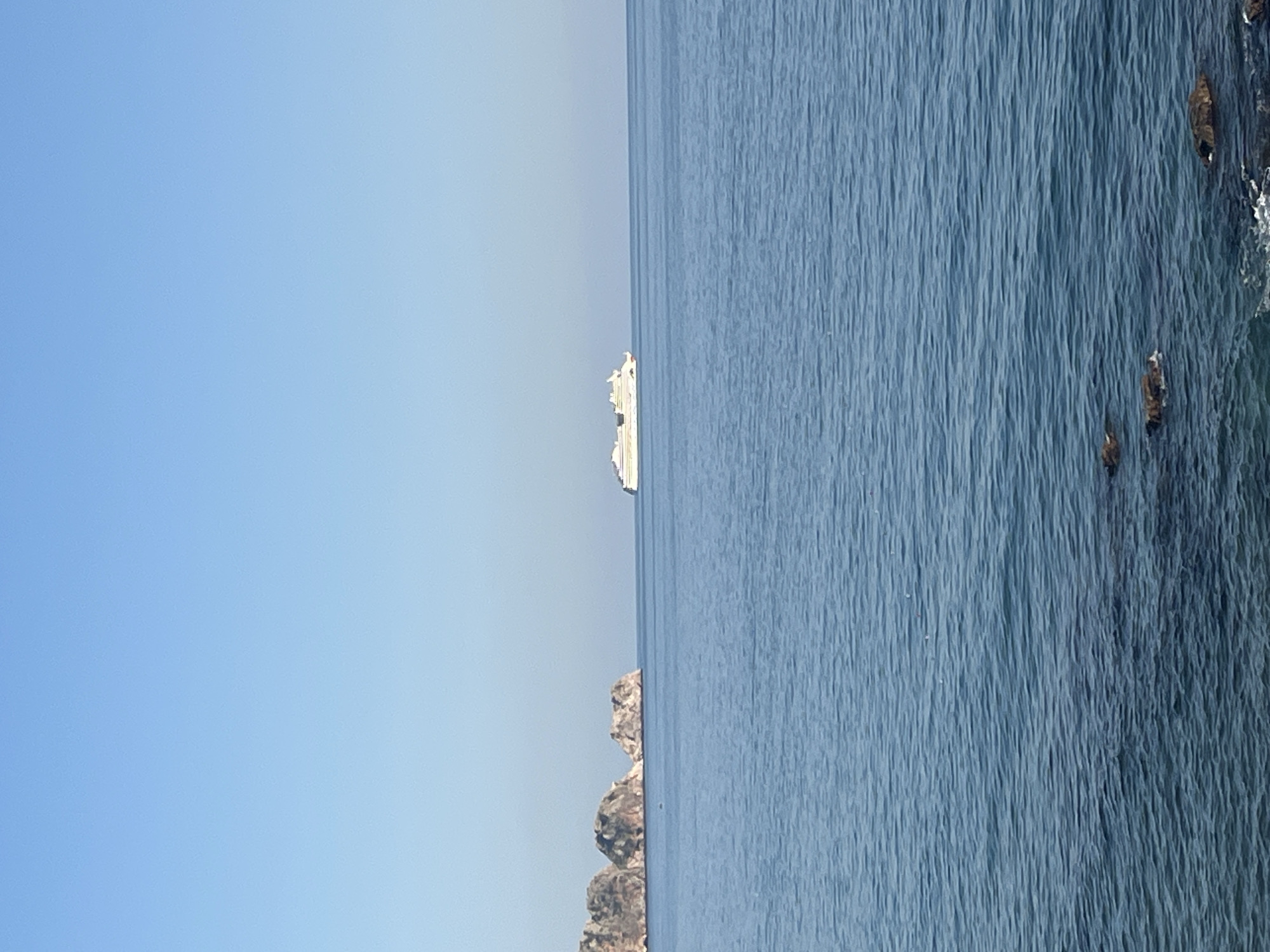 The image presents a serene daytime seascape off the coast of Alanya, Turkey, under a clear and expansive blue sky. The horizon line divides the frame, with the cloudless sky occupying the upper portion and the vast, calm body of water filling the lower half.
In the mid-distance, a prominent white cruise ship or large ferry is visible, sailing across the tranquil blue-grey water. Its clean lines and bright color stand out against the natural backdrop. Closer to the foreground, along the left side of the frame, a rugged, low-lying rocky coastline or a series of small islands emerges from the water. On the far right, darker rock formations are visible near the water's edge, partially submerged, with subtle white foam indicating gentle wave action.
The water's surface is relatively smooth, reflecting the bright light and suggesting calm weather conditions. The overall scene evokes a sense of peace, vastness, and leisure. No people are visible in the photograph, and there is no discernible text. The bright, clear atmosphere suggests a sunny day.