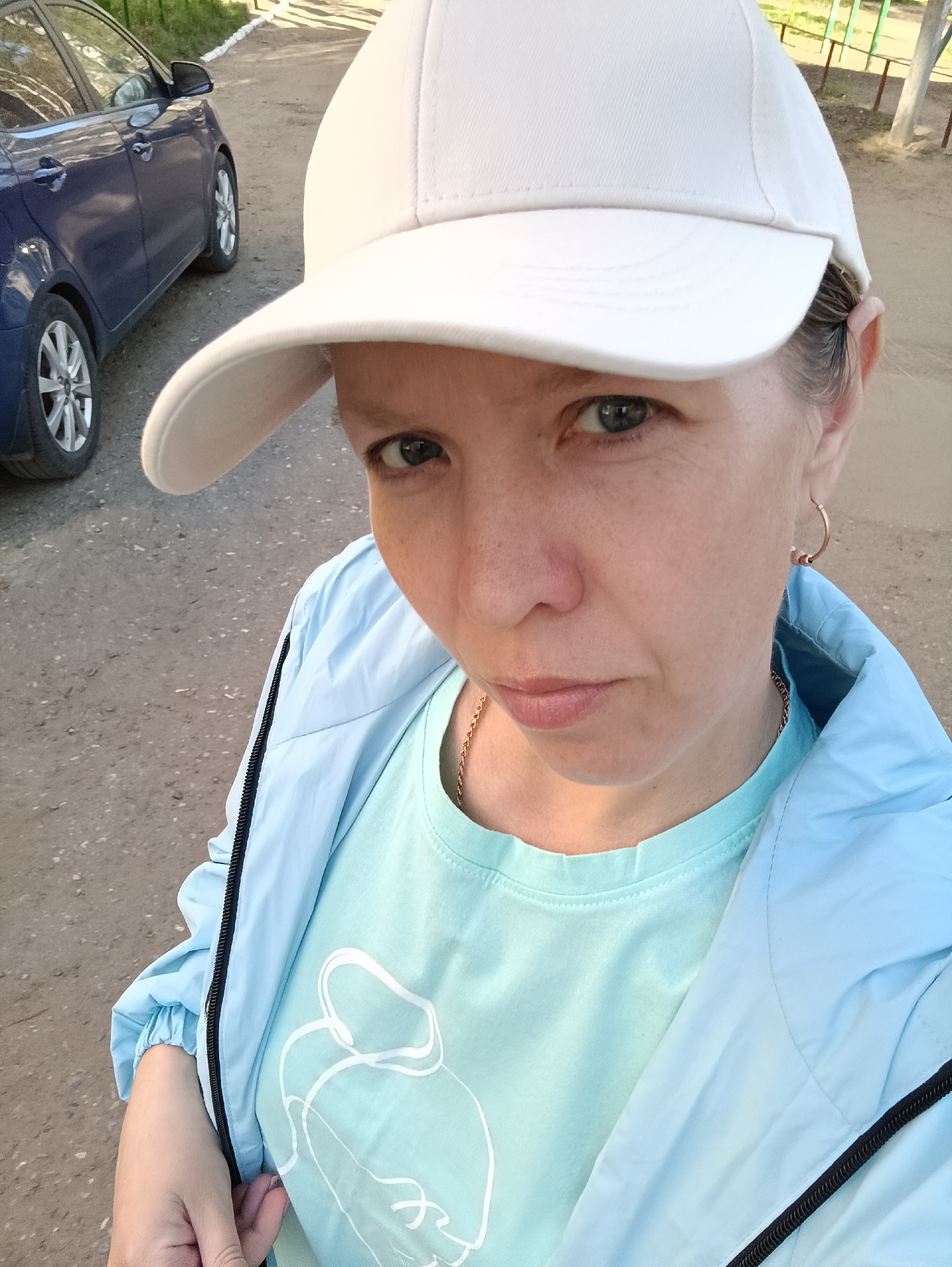 A selfie shows a woman in a white baseball cap, light blue t-shirt with a white design, and a light blue jacket. She has a gold necklace and earrings. The background reveals a paved area, a blue car, and green vegetation, suggesting an urban or suburban setting. Based on additional data, this location is in Chaikovsky, Russia.