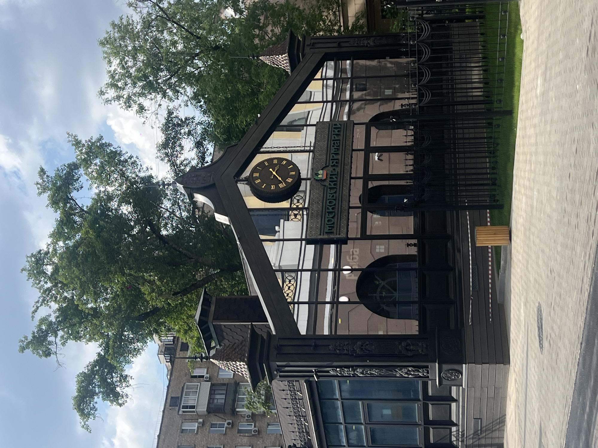 The image shows a clock and sign for "Moskovskiy Yarmarki" in Moscow, Russia. The clock is black with gold Roman numerals and is mounted on a black, triangular structure. The sign is in Russian and features a coat of arms. The structure is in front of a building with large windows and ornate details. A black metal fence is in the foreground. The background includes trees, a cloudy sky, and an apartment building. The scene appears to be outdoors, on a sidewalk or pedestrian area.