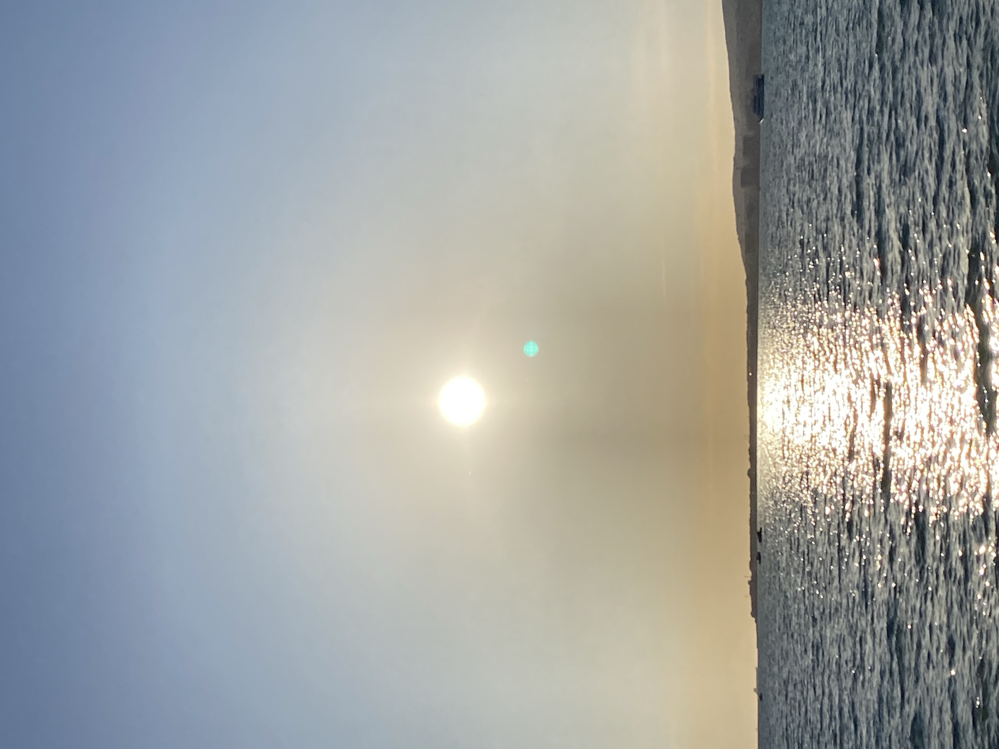 The image depicts a serene natural landscape, likely a coastal view in Gelendzhik, Russia, during either sunrise or sunset. The sky occupies the upper portion of the frame, transitioning from a soft, light blue at the top left to a warm, hazy golden-white around the sun. A bright, circular sun is prominently featured towards the center-left, emanating strong light and a subtle lens flare that appears as a greenish-blue dot to its right.
Below the sky, a vast body of water, presumably the Black Sea, stretches across the lower right. The water's surface is characterized by numerous ripples or small waves, which intensely reflect the sunlight, creating a shimmering path of bright white and golden sparkles. A thin, muted strip of land or coastline is visible along the horizon line, separating the water from the sky, appearing as a dark, indistinct silhouette against the bright backdrop.
The time of day is clearly indicated by the low position of the sun and the golden hues, suggesting either a tranquil morning sunrise or an evening sunset. The weather appears clear with some atmospheric haze, particularly around the sun, which diffuses the light and contributes to the warm, glowing ambiance. No people, objects other than the natural elements, or visible text are present in the image.