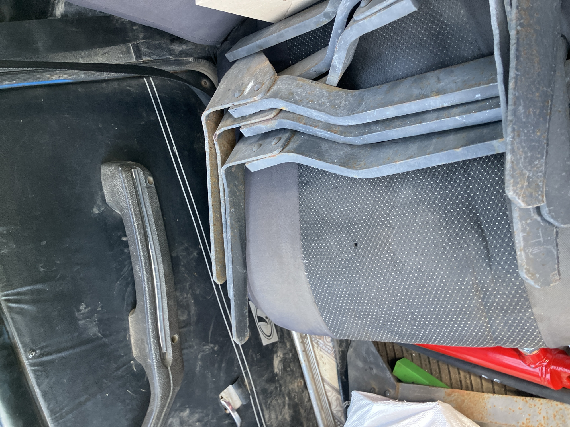 In the city of Molodizhne, Ukraine, this image captures the interior of a vehicle, likely a car or van. The focus is on the passenger seat area, which is mostly covered by a stack of dark grey, metal brackets. These brackets are bent at right angles and appear to be industrial or automotive parts, some showing signs of rust.
The backdrop consists of the car's interior: a black door panel with a textured, worn surface and a chrome-accented handle. A single white circular sticker with a black logo is visible on the door panel. A portion of the car's seat, upholstered in dark grey fabric with a subtle dot pattern, is also visible. In the lower left corner, a white bag and a bright red cylindrical object are partially visible, along with a bright green object. The overall impression is one of storage or transport of tools or parts within the vehicle.