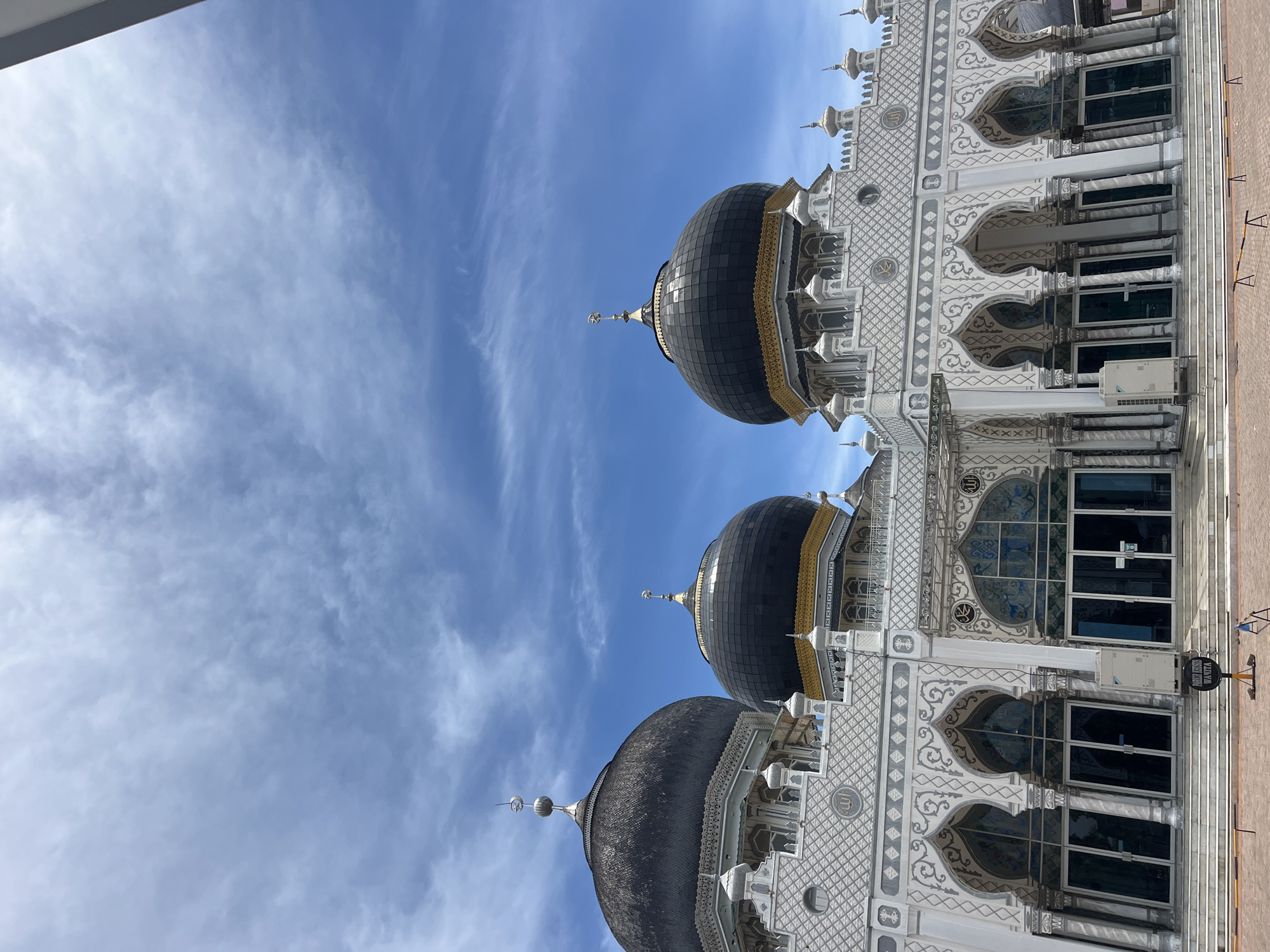 The image shows a low-angle view of the Baiturrahman Grand Mosque in Keudekruenggeukueh, Indonesia. Three dark-colored domes with golden accents are prominently featured against a blue sky with wispy clouds. The mosque's facade is intricately designed with white and gray geometric patterns, arches, columns, and several windows. The architecture appears well-maintained. Part of a sidewalk is visible on the right edge of the frame.