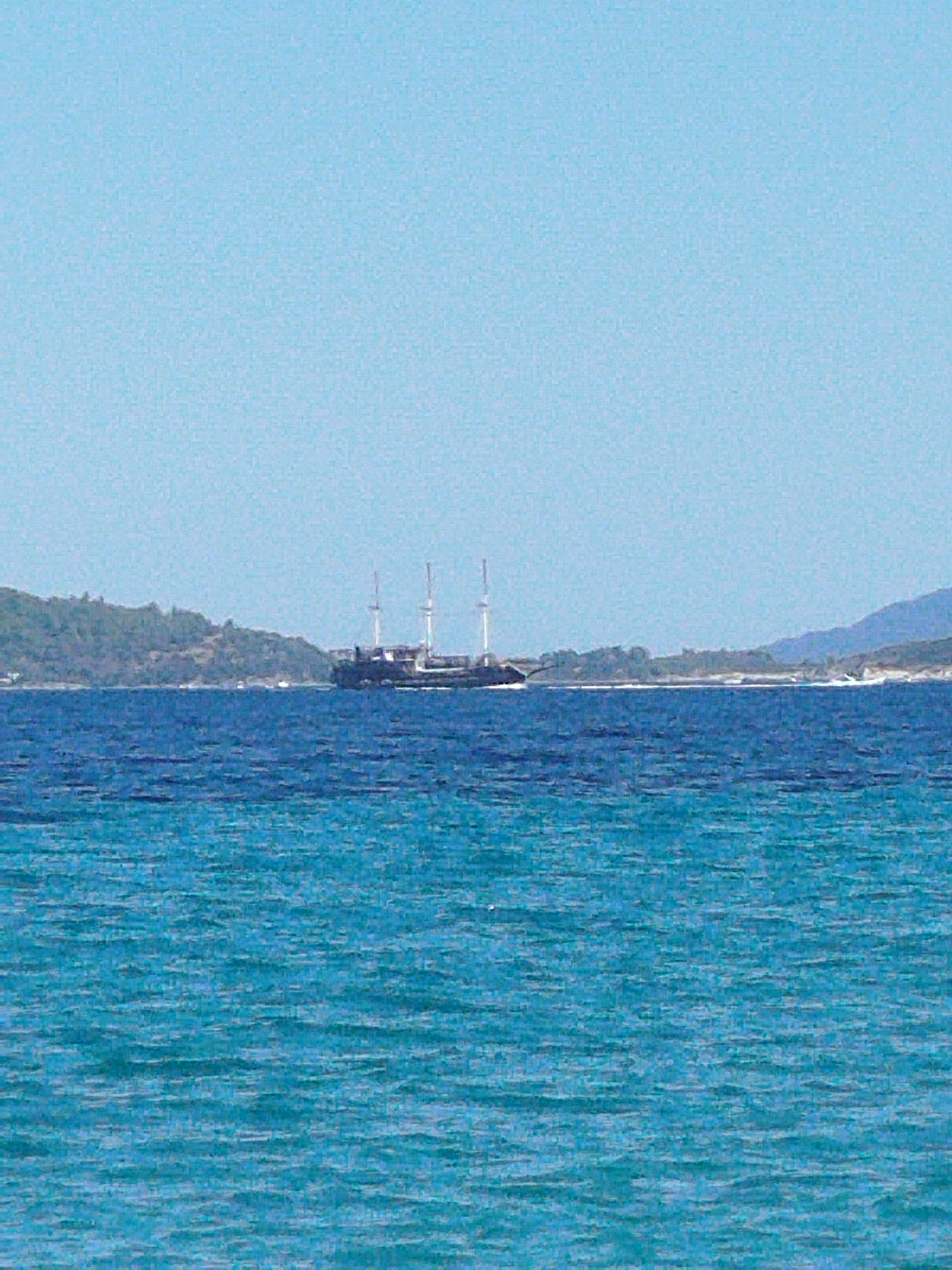 The image shows a wide-angle view of the sea off the coast of Sithonia Municipal Unit, Greece. The water in the foreground is a bright turquoise with small waves, transitioning to a deeper blue in the midground. In the distance, a large sailing ship with three masts is visible against a backdrop of green, tree-covered hills and a hazy sky. The ship appears to be sailing towards the right of the frame. The overall scene depicts a calm day at sea.