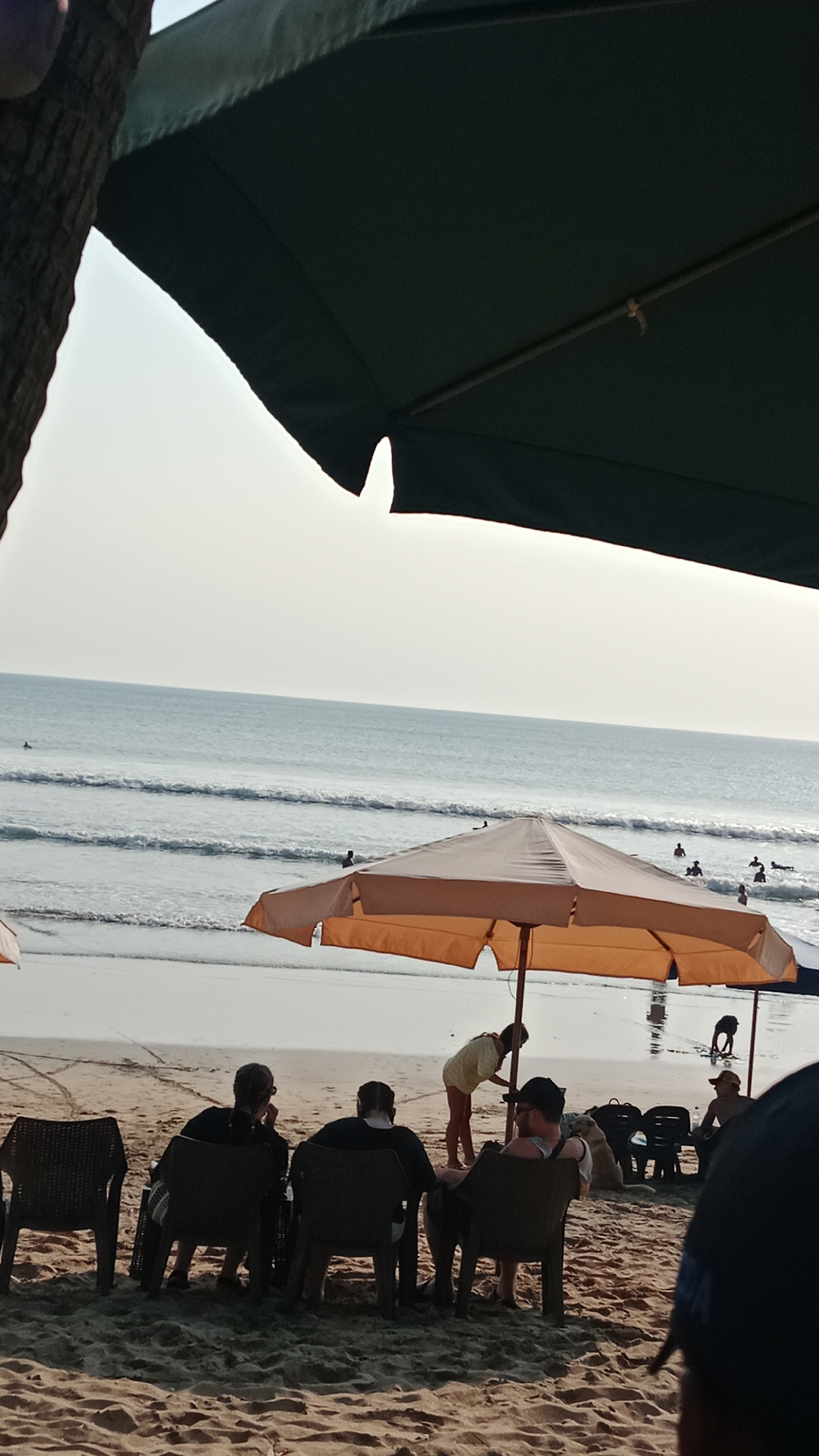 This is a vertical, medium-distance, eye-level shot of a beach scene in Kuta, Indonesia, likely taken during the late afternoon. The foreground is dominated by sand, with several dark, plastic beach chairs arranged on it. In the immediate foreground, three people are seated facing away from the camera, appearing to be observing the ocean. One person on the far left is wearing a dark shirt, the middle person is wearing a dark shirt and possibly a patterned bag on their lap, and the person on the right is wearing a dark shirt and cap.
Behind these seated figures, a large, light orange beach umbrella provides shade. A woman in a yellow top and light-colored shorts is standing near the umbrella, bending over and appearing to be attending to something on the ground or interacting with a person out of frame. Further back, on the beach and closer to the water, more beach chairs are visible, some occupied by people.
The middle ground features the ocean, with gentle waves rolling towards the shore. Several figures are visible in the water, some appearing to be swimming or surfing. The water is a light blue-grey color, and the sky is a pale, hazy blue, suggesting the sun is low in the sky or obscured by haze. The overall atmosphere is relaxed and recreational.