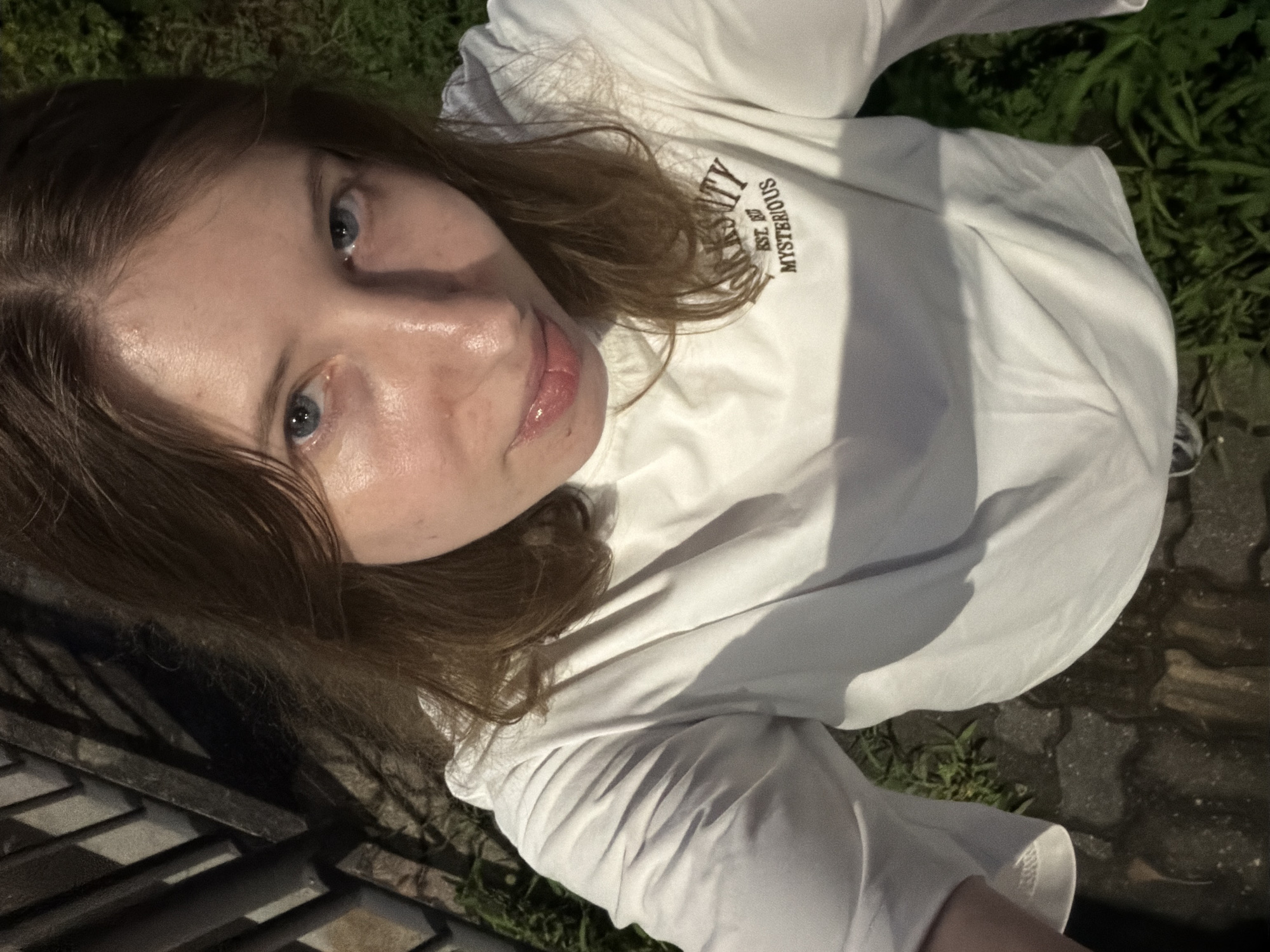 This is a first-person perspective photograph of a young person with light skin and brown hair, wearing a white long-sleeved t-shirt. The t-shirt has black text on it that reads "MYSTERIOUS EST. 1997". The subject's face is looking upwards towards the camera, with blue eyes and slightly parted lips. The lighting suggests it is daytime, possibly late afternoon or early evening, with dappled shadows from an unseen light source. The background consists of green foliage and a paved area with interlocking stones, suggestive of an outdoor setting such as a park or garden. There appears to be a metal railing or fence in the lower left portion of the image. The overall mood is casual and informal.