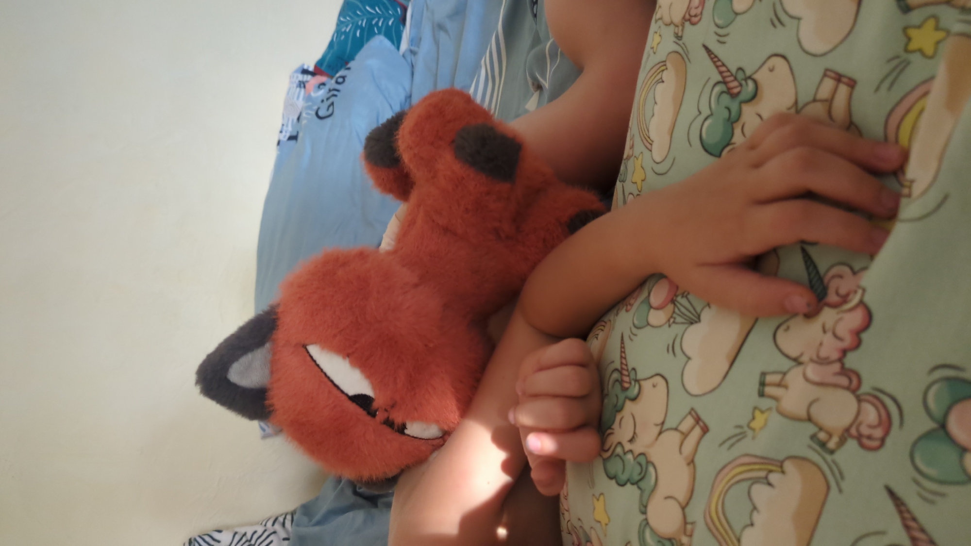An overhead, close-up shot shows a child's arm resting on a light blue pillow with a whimsical unicorn pattern. The child's hand is relaxed, with fingers slightly curled, and a plush fox toy is tucked behind their arm, its orange fur and black ears visible. The background consists of rumpled blue bedding and a plain, off-white wall. The lighting suggests a sunlit indoor environment. This image was taken in Chișinău, Moldova.