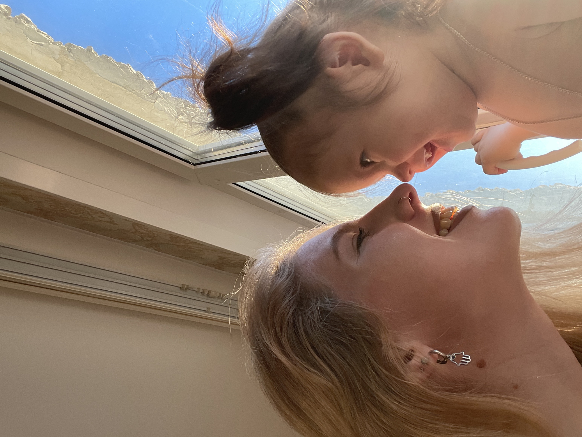 A low-angle shot depicts a woman looking up at a baby. The woman has fair skin, blonde hair, and is wearing an earring shaped like a hand. The baby, positioned above the woman, has dark hair pulled into a bun. They are both situated indoors, with a window and what appears to be the ceiling visible in the background. The location in the city of Izhevsk, Russia, cannot be determined from the visual information provided.