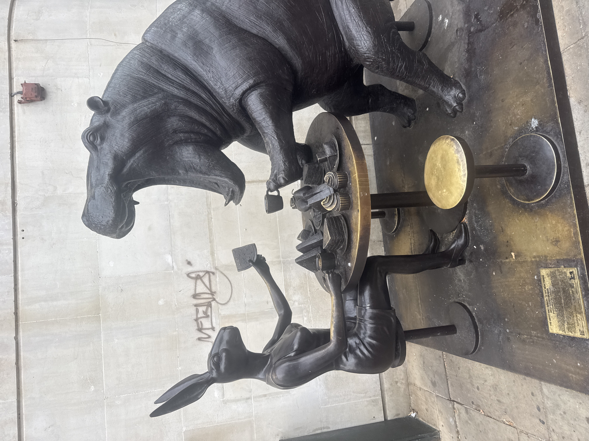 In London, United Kingdom, a bronze sculpture depicts a scene of two figures at a table. A rabbit woman, dressed in a black dress and heels, sits on a stool to the left of a round table laden with cupcakes and slices of cake. She holds a square object, possibly a card or a small plate. Opposite her, a hippopotamus sculpture with its mouth wide open is seated, holding a teacup in its left hand. The table appears to be made of dark metal with a gold-colored rim. A smaller gold-colored stool is positioned in front of the hippo. The sculpture is situated on a raised metal base, which has a plaque at the front. Behind the sculpture, a light-colored brick wall is visible, with some graffiti marks present. The ground in front of the base is paved with grey flagstones. The overall scene appears to be an outdoor installation.