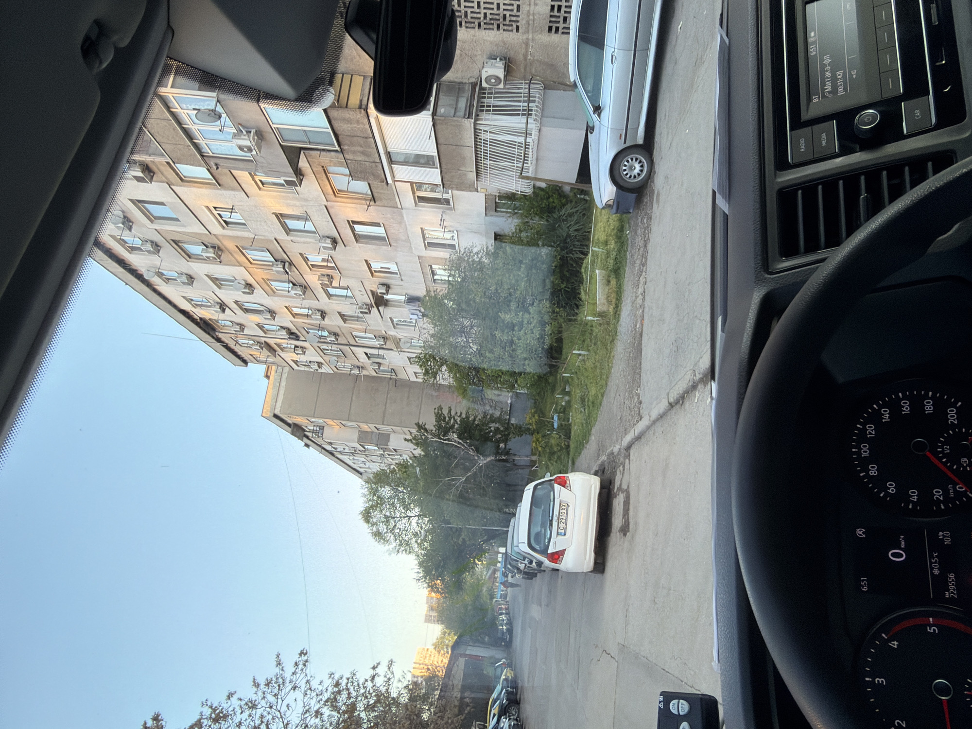 Captured from the interior of a parked vehicle, this image presents a view of a residential street in an urban setting in the city of Unknown, Unknown. The prominent feature of the external environment is a tall apartment building with numerous windows and air conditioning units affixed to its exterior. Adjacent to the building, along the street, a row of parked vehicles extends into the distance, including a white car directly in front of the vehicle the image is taken from. The street is lined with greenery, including trees and grass.
Inside the vehicle, part of the dashboard and steering wheel are visible. The dashboard features a radio/media console with an active display screen, while the instrument cluster indicates the vehicle is stationary, with a temperature reading of 0.5 degrees Celsius. The odometer displays a reading of 229556. The time is 6:51.