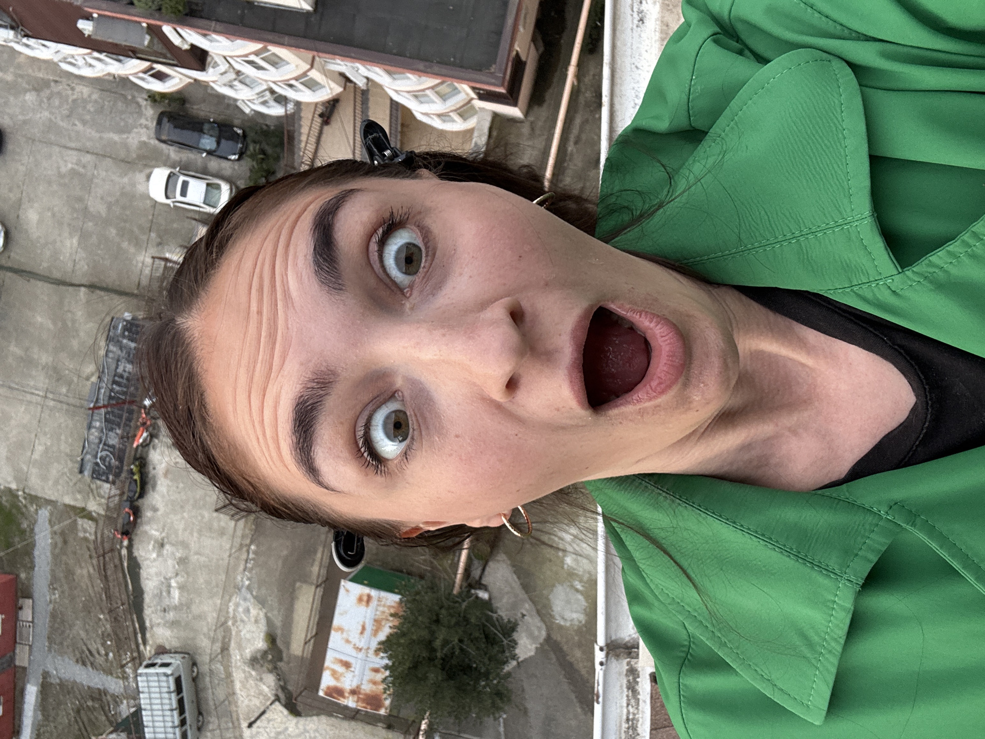A selfie taken from a high-angle perspective shows a woman with a surprised expression, wide eyes, and open mouth. She is wearing a bright green jacket and a black top. Her brown hair is pulled back with clips. She has light skin, arched eyebrows, and is wearing hoop earrings.
The background suggests she is on a high balcony or rooftop in an urban area. Below, buildings and streets are visible. Parked cars, a small tree, and other urban elements are identifiable.
The image was likely taken in an urban area with multi-story buildings, possibly in Sochi, Russia, based on the style of architecture and general environment.