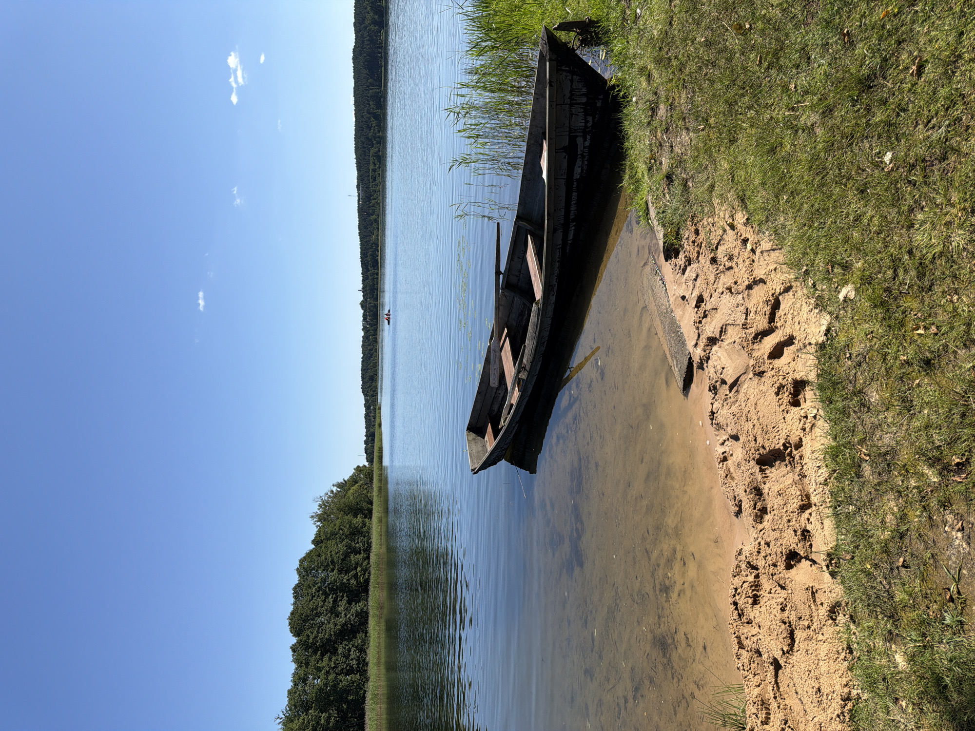 An eye-level, medium shot captures a serene lake scene under a clear blue sky. In the foreground, a dark wooden rowboat rests partially on a sandy shore, its interior visible with wooden seats. The lake's surface is calm, reflecting the sky and the dense green treeline that borders the opposite bank. Reeds grow along the right edge of the water, near where the boat is beached. In the distance, two figures can be seen in a small boat on the lake. The overall impression is one of peaceful solitude in a natural setting.