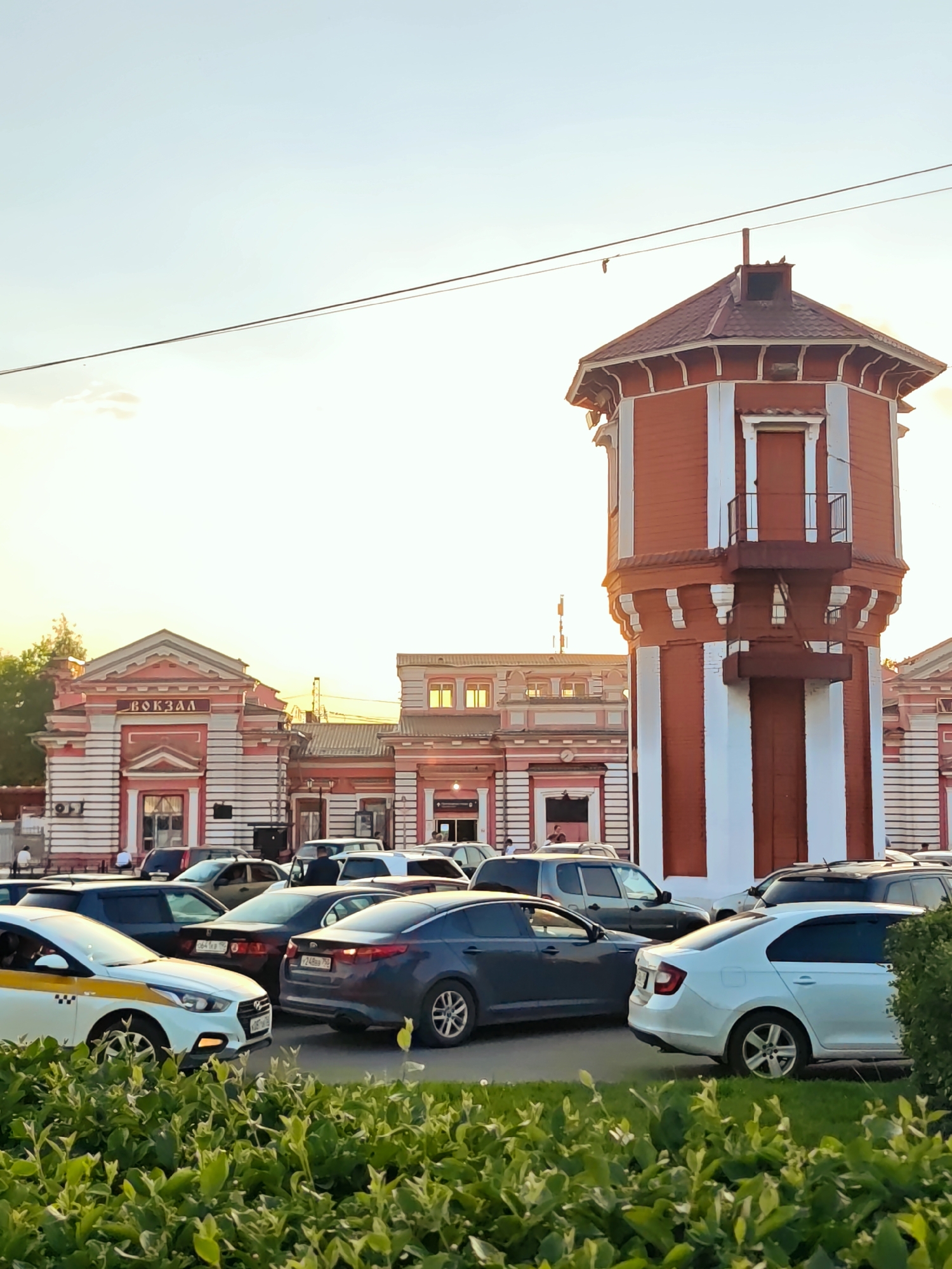 The image depicts a scene in Dmitrov, Russia, featuring the Dmitrov railway station and its distinctive water tower. The railway station, with its red and white facade and the Russian word "Вокзал" (Vokzal, meaning "Station") above the entrance, is partially visible behind a parking lot filled with various cars, including a taxi. The red and white water tower stands prominently to the right of the station building. Lush green bushes occupy the foreground. The lighting suggests either a sunrise or sunset, casting a warm glow over the scene. Several people can be seen in the station area. Power lines are visible against the sky.