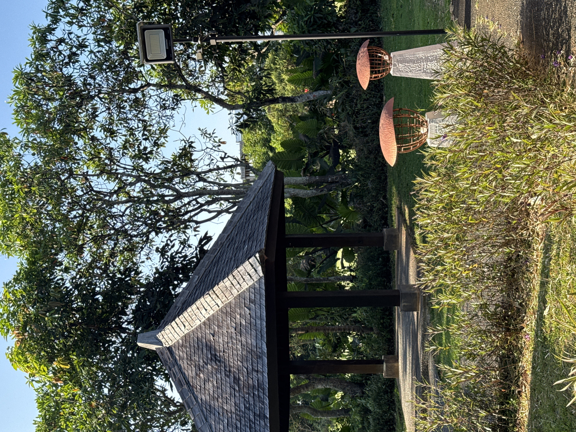 The scene depicts an outdoor setting in Kuta Selatan, Indonesia. Dominating the view is a wooden gazebo with a tiled roof, set amidst lush greenery. To the right, two decorative sculptures, consisting of copper-colored half-spheres atop white pedestals, flank a paved pathway. A bush with green foliage is also prominent. In the background, a modern streetlight stands tall. The trees suggest a tropical climate, which is typical of the area.