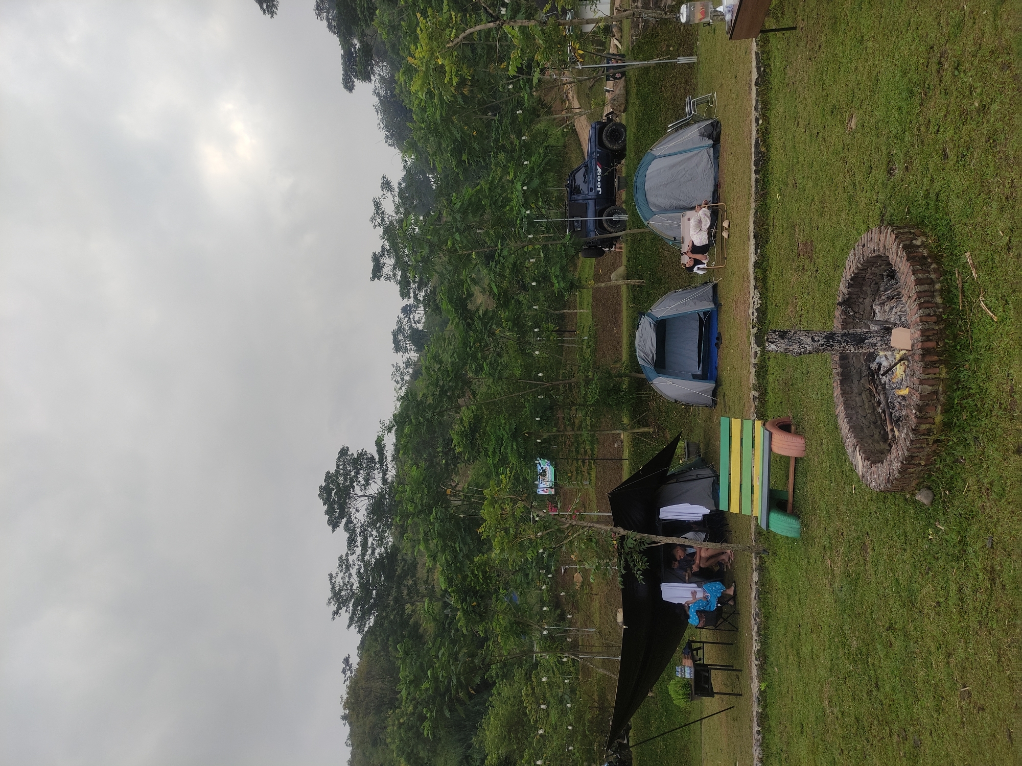The image depicts a campsite in Babakan Madang, Indonesia, on an overcast day. The site is situated on a grassy area near a wooded hillside. Three tents are visible: two are dome-style, and one has a dark tarp structure erected over it. People are present near the tents, but their actions are unclear. A blue Jeep is parked near the tents. A brick-lined fire pit contains charred wood. A multi-colored bench made of wood and tires sits between the tarp-covered tent and the fire pit. String lights hang from the trees. The hillside is covered in dense trees. The sky is uniformly grey with no visible sun.