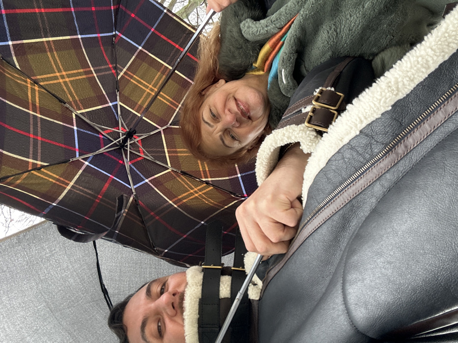 The image displays two individuals, a male and a female, situated outdoors in Sofia, Bulgaria, captured from a low angle. On the left, a male subject with dark hair and a mustache is visible, smiling slightly. He is wearing a dark, possibly leather or synthetic, jacket featuring a prominent cream-colored shearling collar and lining, with dark brown straps and brass-colored buckles visible. He holds the handle of a solid gray umbrella, which partially extends into the upper left of the frame. On the right, a female subject with reddish-brown hair is seen smiling towards the camera. She wears a green, possibly textured or fuzzy, outer garment and a multi-colored scarf displaying blue, orange, and yellow tones. She holds the handle of a dark plaid umbrella, patterned with intersecting brown, red, blue, and yellow lines, positioned open above her. The background is blurred, showing indistinct tree branches against a light, overcast sky, suggesting an outdoor environment likely experiencing or having recently experienced precipitation.