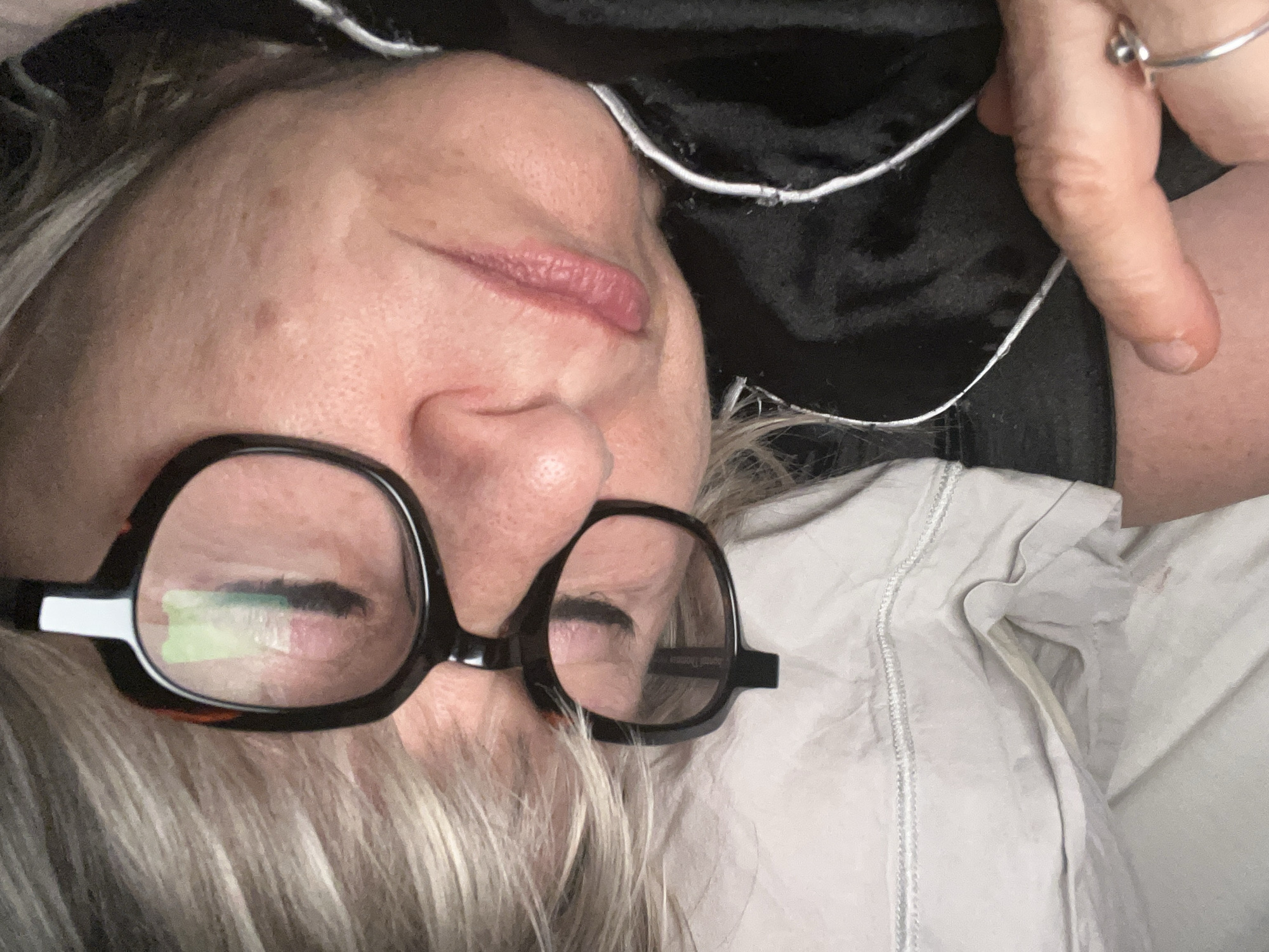 A middle-aged woman is lying on her side, likely on a bed. She is wearing glasses and has gray hair. She has on what appears to be a dark-colored garment with a white trim at the neckline. Her hand is raised near her face, with a ring visible on one of her fingers. She is wearing light-colored pajamas.
There is no information available about the location being in Chambœuf, France.