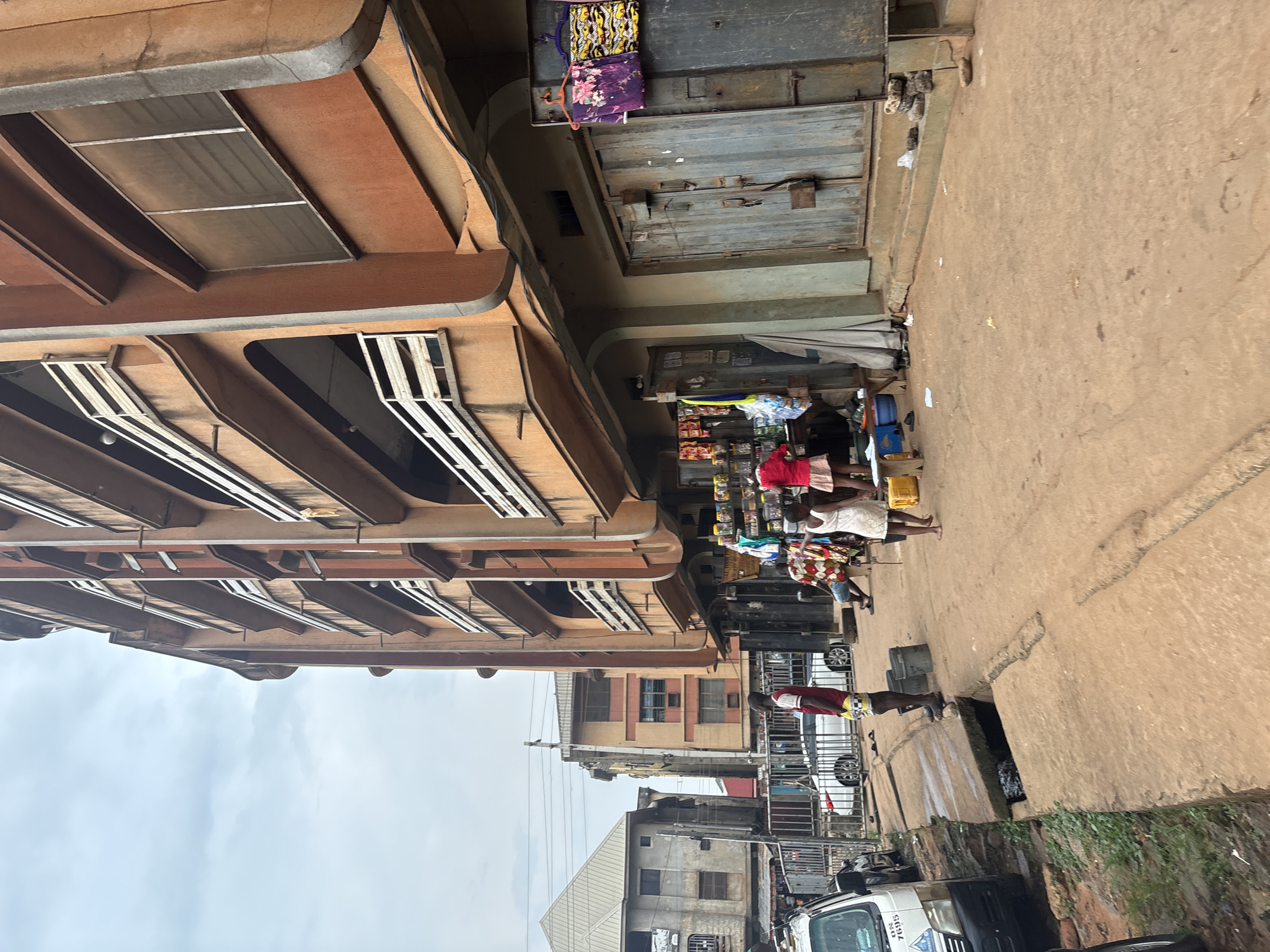This image depicts a street scene in Onitsha, Nigeria, characterized by a multi-story building with an orange-brown facade and balconies featuring white railings. The building appears to house shops on its ground floor, indicated by closed metal doors and displays of goods. Several people are present, including a man standing near a drain, a woman in a red top and skirt, and two girls in white dresses. The ground is a dirt or concrete surface, with a visible drainage channel running along the street. A white van is parked on the left side of the image. The sky is overcast, suggesting a cloudy day. The overall impression is of a busy urban environment with commercial activity.
