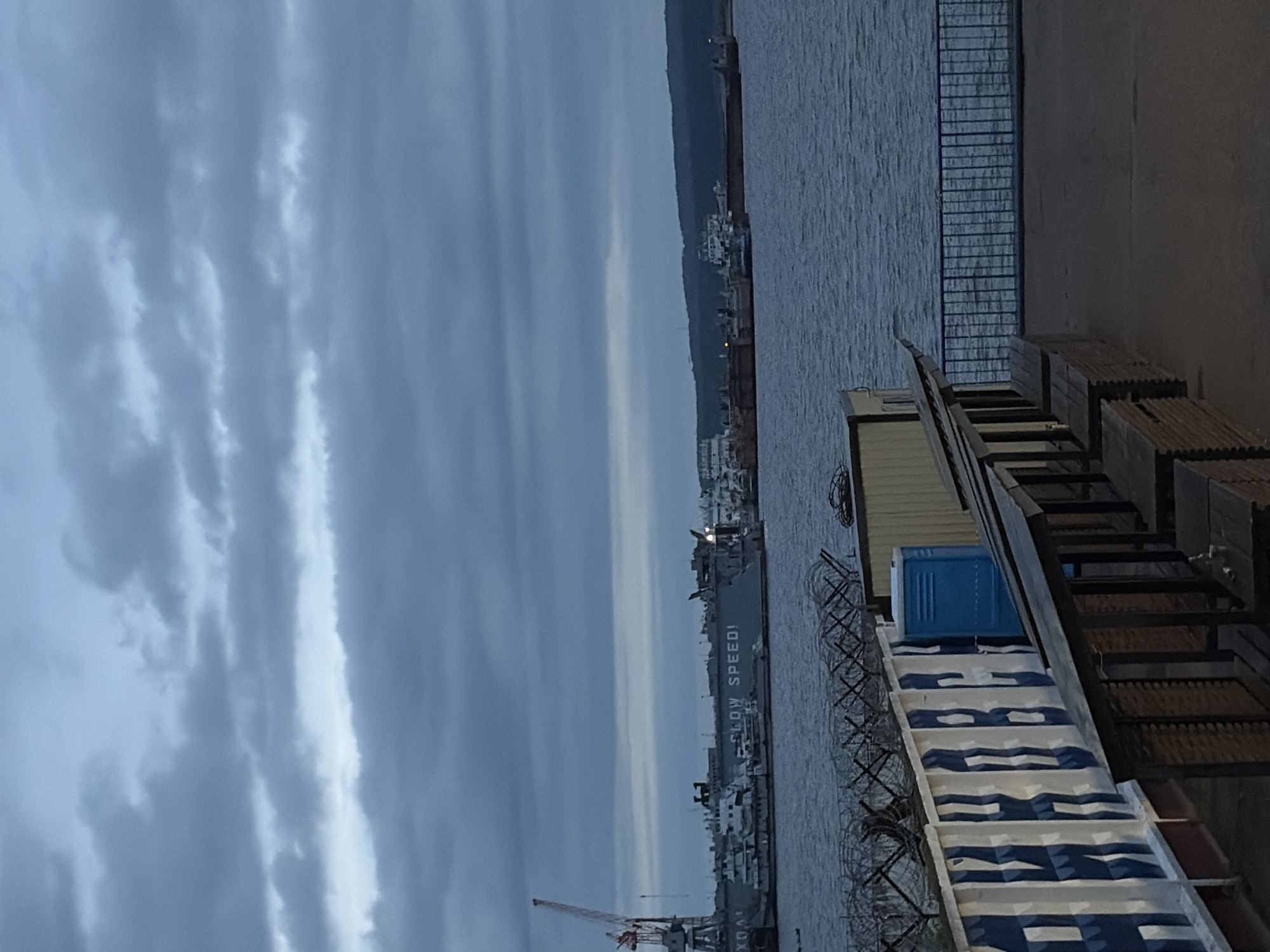 The image depicts an overcast waterfront or port scene. A vast, dark grey-blue sky filled with dense clouds covers the upper half of the frame, with a slightly lighter horizontal band visible on the horizon. A body of water, dark blue-grey with subtle ripples, occupies the middle ground.
In the mid-distance, a busy port area is visible. A large, grey vessel, possibly a dry dock or cargo ship, is prominent, displaying the white capitalized text "SLOW SPEED!" on its side. To the left of this vessel, a red and white port crane stands tall. Multiple other ships and barges are docked or moored across the water, and a dark green, undulating landmass forms the distant background.
The foreground features a concrete or paved platform. On the left side, a low wall or structure is painted with stylized blue and white patterns, resembling Cyrillic letters, partially spelling what appears to be "АПИНОВА". Behind this structure, multiple coils of concertina wire are arranged along what seems to be a barrier. A blue portable toilet is positioned next to the patterned structure and behind the wire. On the right side of the foreground, a row of simple wooden benches faces the water, extending from a light-colored building partially visible on the far right. A blue metal railing separates the platform from the water's edge.
The overall lighting is dim and even, consistent with an heavily overcast day or twilight. No people are visible in the scene.