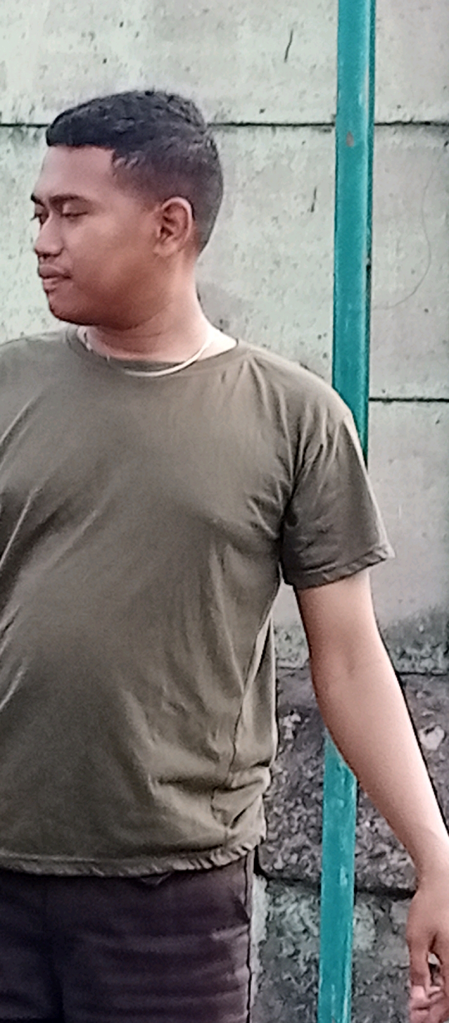 A young man of East Asian descent with dark hair and brown skin stands in front of a grey, textured wall. He is wearing a short-sleeved, olive-green t-shirt and dark-colored trousers. A thin, silver necklace is visible around his neck. To his right, a vertical, teal-colored pole is present. The background appears to be an outdoor setting, possibly a street or an alley, in Wadungasri, Indonesia, suggested by the wall's texture and the overall lighting. The man is looking to his left, with his right arm extended downwards.