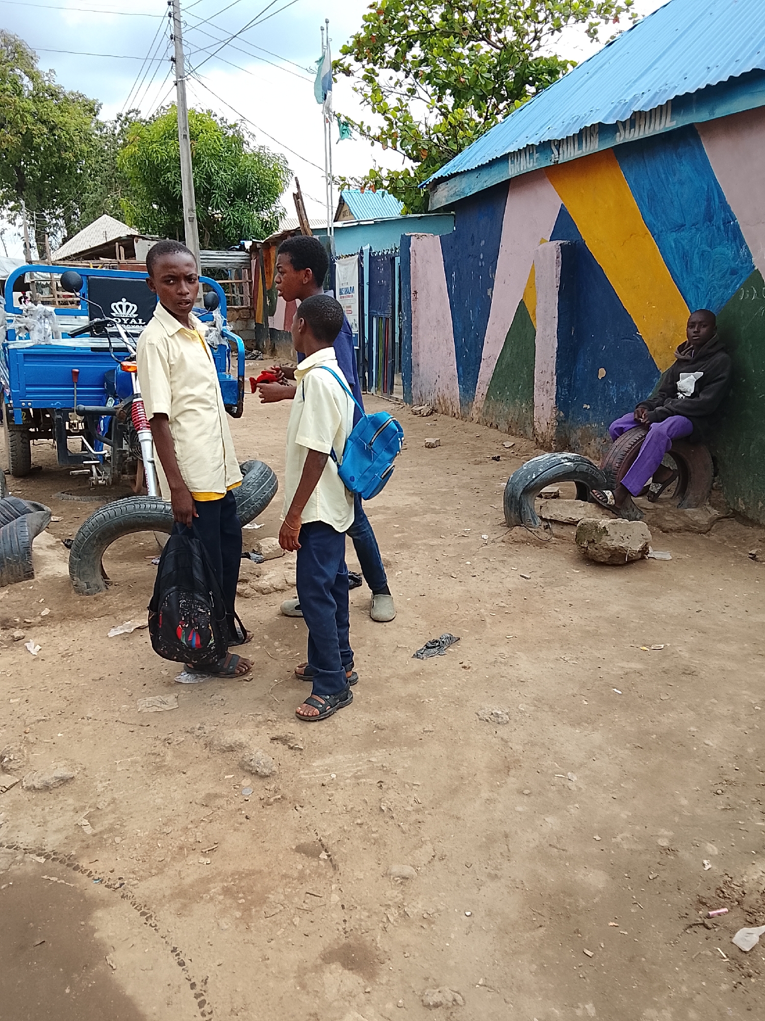 The scene appears to be a dusty street in a low-income area. In the foreground, three young boys are standing. Two of the boys are wearing similar light yellow shirts and dark pants. One is holding a black backpack, and the other has a blue backpack. A third boy is wearing a blue shirt. Behind the boys is a blue three-wheeled vehicle. To the right of the boys, there is a building with a blue roof and a wall painted with colorful geometric shapes in shades of blue, yellow, pink, and green. A young person wearing a black hoodie and purple pants is sitting on a tire leaning against the painted wall. There are tires scattered around on the dirt ground. It is daytime under a cloudy sky. There is no information that identifies the location as the city of Abuja, Nigeria.