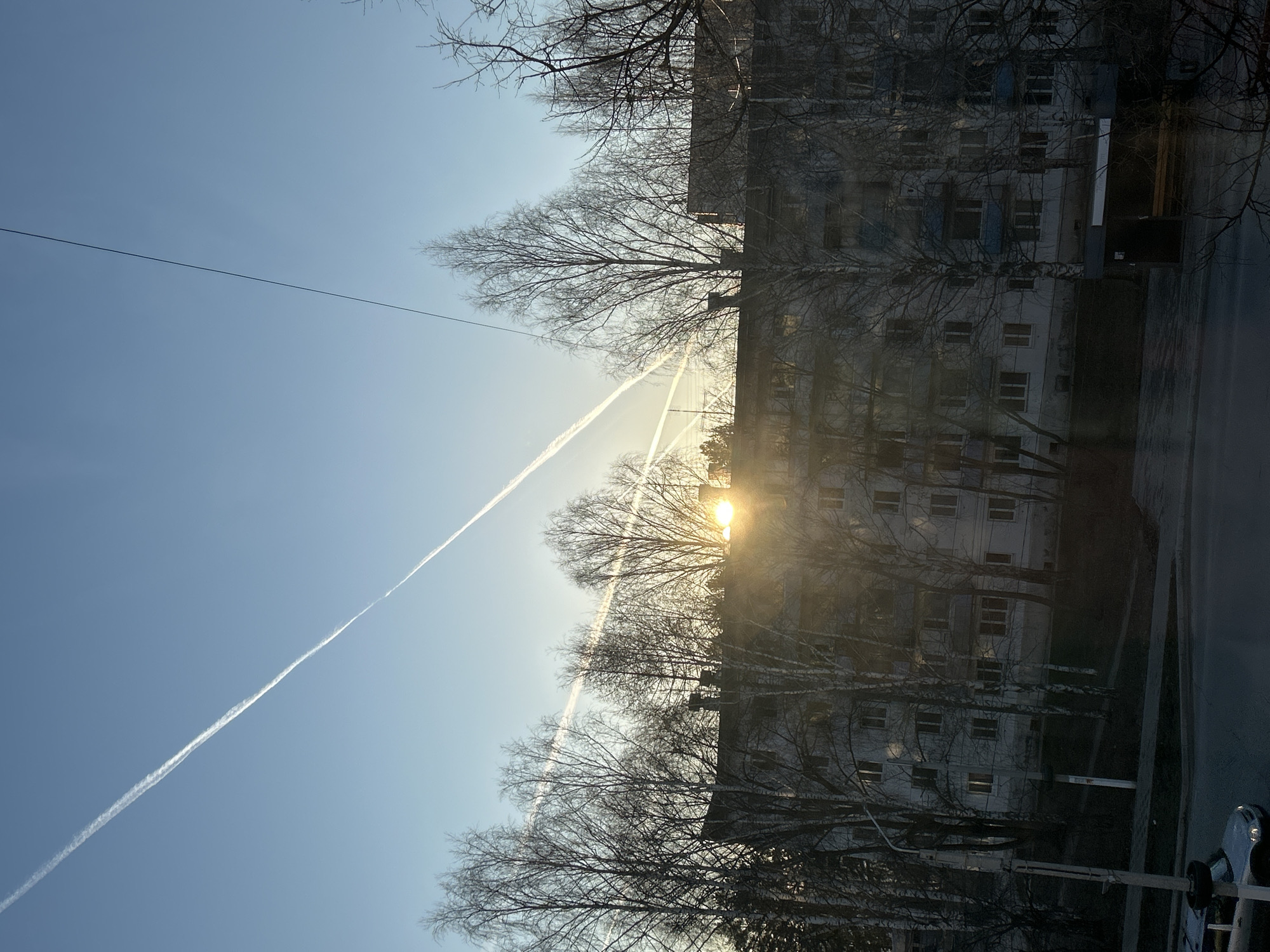 The image shows an urban landscape in Chaikovsky, Russia at what appears to be dawn or dusk. The sky is light blue with visible contrails from aircraft. An apartment building stands to the right of the frame, obscured by bare trees. The sun shines behind the trees, creating a bright glare. A utility wire stretches across the upper left portion of the sky. A sidewalk and road are visible in the lower right, with a portion of a vehicle appearing in the very bottom corner. No people are visible.