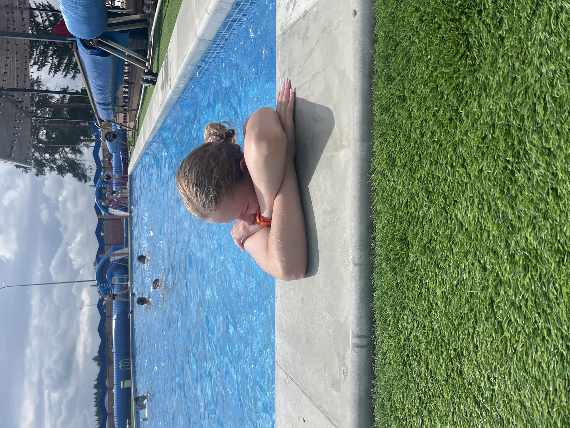 A young girl with blonde hair pulled into a bun rests her head on her knees, her arms wrapped around her legs. She is partially submerged in a bright blue swimming pool, with water droplets on her arms. The pool has a tiled bottom and appears to be part of a water park, with blue slides visible in the background. Green artificial turf lines the edge of the pool. The sky is cloudy, suggesting it could be a partly cloudy day. Other swimmers can be seen in the pool in the distance. The overall scene suggests a leisurely day at a water park. There is no visible text in the image.