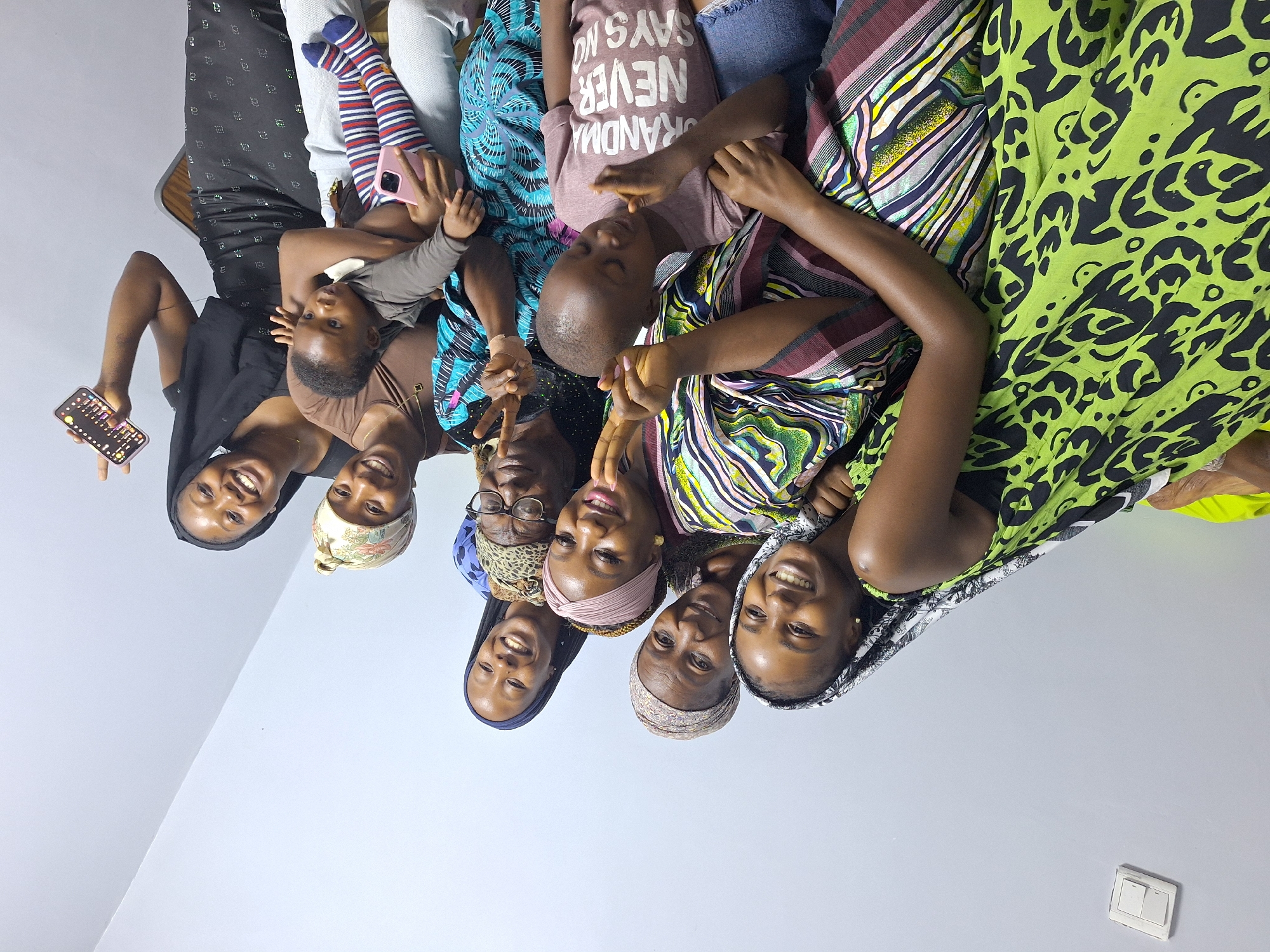 A group of nine individuals, including adults and two young children, are posing for a photograph in an indoor setting, likely a home, in Sabon Tasha, Nigeria. The group is arranged in a semi-circle, with some individuals standing and others seated or holding children.
In the foreground, a young boy wearing a dusty rose t-shirt with white lettering that reads "GRANDMA NEVER SAYS NO" is looking down at his hands. To his right, a baby is being held by a woman in the background. The baby is wearing striped leggings and looking to the left.
The adults are dressed in a variety of colorful clothing, including patterned dresses, headscarves, and blouses. Several individuals are smiling and making peace signs with their fingers. One woman on the far right is holding up a smartphone, which appears to be displaying colorful icons.
The background consists of a plain, light-colored wall and a portion of a light switch on the upper left side. The lighting suggests an indoor environment, possibly illuminated by overhead lights or natural light from a nearby window. The overall atmosphere appears to be joyful and familial.