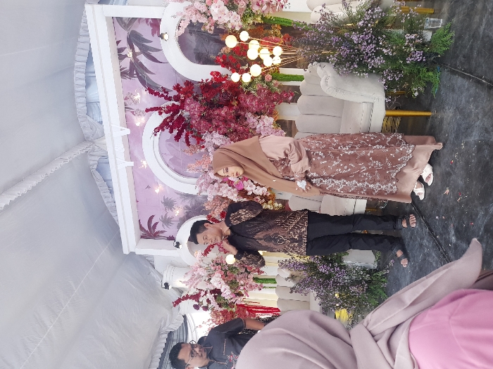 This image captures a formal event, likely a wedding or engagement ceremony, taking place under a white tent in Pacul, Indonesia.
**Scene:** The central focus is an elaborately decorated stage area. The background features a large white framed structure with arched cutouts, showcasing a soft pink and purple mural depicting dark silhouettes of palm trees against a sky-like motif, accented with small embedded lights. Vibrant floral arrangements, dominated by shades of red, pink, and purple, densely adorn the backdrop and frame the central section. Two elegant, light-colored (cream or off-white) sofas with gold-colored legs are positioned in front of the backdrop, flanked by clusters of purple flowers. The ground is covered with a dark, possibly black, surface. Overhead, the white fabric of the tent ceiling is visible.
**People:**
* **Central Man:** Standing slightly to the left of the center, a man with dark hair, appearing to be in his 30s or 40s, wears a dark brown batik-patterned long-sleeved shirt, black trousers, and dark sandals. His right hand is raised to the side of his head or ear.
* **Central Woman:** Standing to the man's right, a woman, also appearing to be in her 30s, wears a light brown hijab, a long-sleeved light brown dress, and a darker brown overlay adorned with white patterns. She wears white open-toed sandals.
* **Foreground Figures:**
* On the far left, partially visible, a man wearing glasses and a dark, patterned short-sleeved shirt is observed looking towards the central man.
* In the immediate foreground on the left, the back of a person wearing a light pink top and a light brown hijab is visible, facing towards the stage.
**Objects and Actions:** The central man appears to be speaking or adjusting something near his ear. The central woman stands beside him, observing the scene. Decorative light fixtures with spherical bulbs are visible on the right side of the stage. The overall impression is a staged event with guests observing the central figures.
