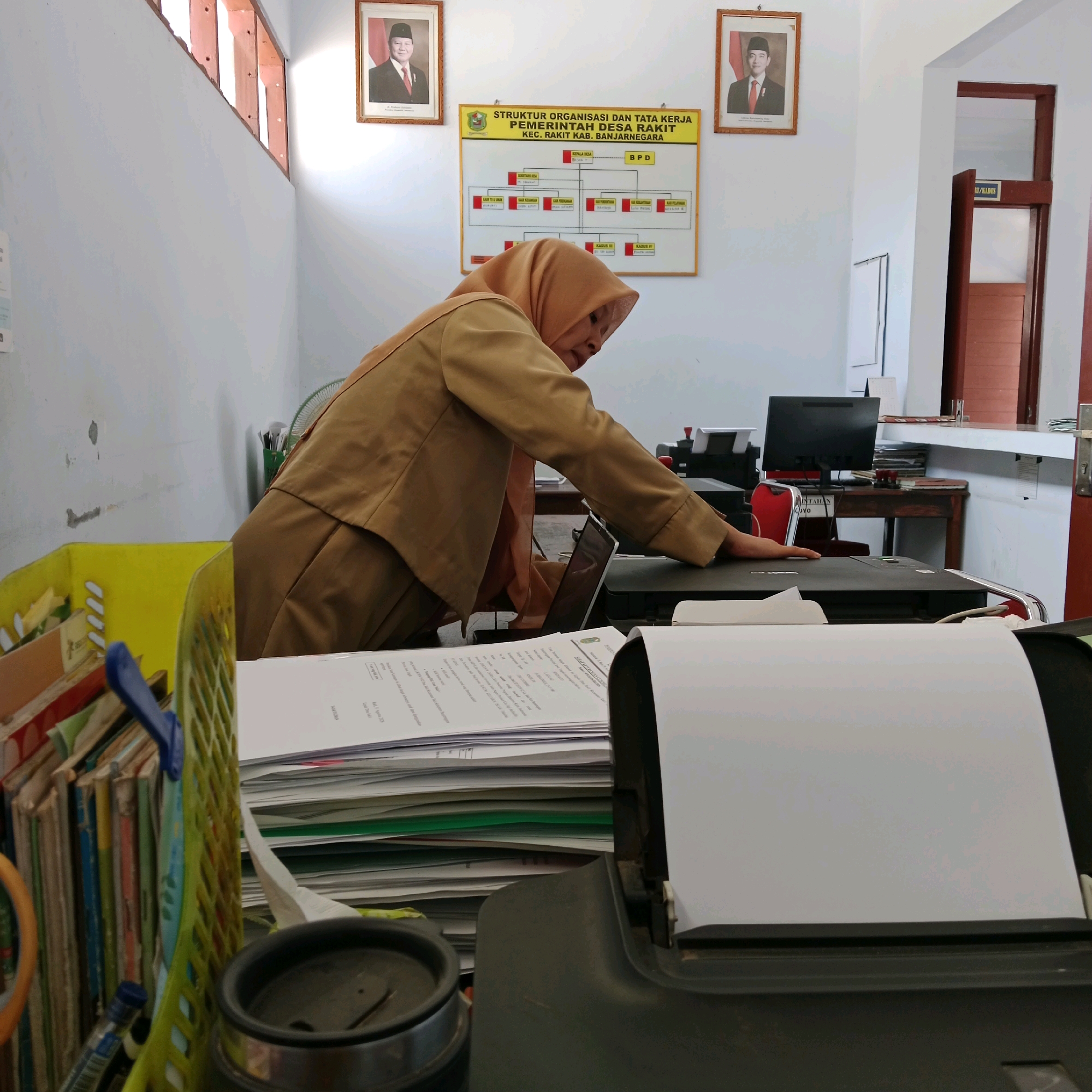 The image depicts the interior of an office, likely in the village of Rakit, Banjarnegara Regency, Indonesia. A woman, dressed in a tan uniform with a matching headscarf, is leaning over a printer, her right hand on the device. She appears to be operating or interacting with the printer, possibly preparing to print documents.
The office environment is evident from the items visible. On the wall behind the woman, there is an organizational chart titled "STRUKTUR ORGANISASI DAN TATA KERJA PEMERINTAH DESA RAKIT KEC. RAKIT KAB. BANJARNEGARA". Portraits of officials are also displayed on the wall.
In the foreground, a large stack of papers and a black printer are prominent. To the left, a yellow plastic basket is filled with books and files. A computer monitor and desk are visible in the background, suggesting a typical office setup. The overall impression is one of a working administrative office.