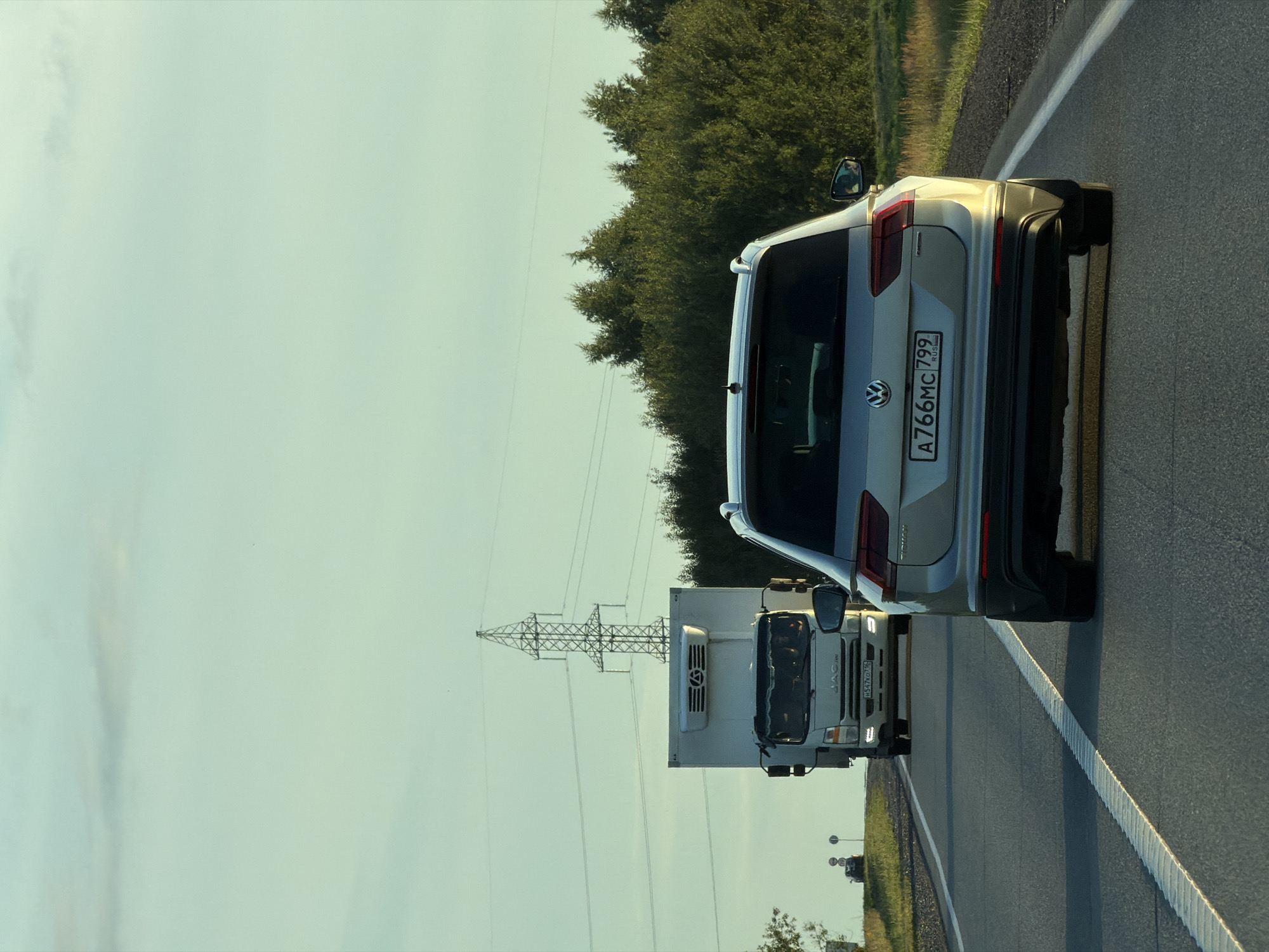 A medium shot captures a silver Volkswagen Tiguan driving on a highway in the city of Obukhovo, Russia, during daylight. The car's rear is visible, showing its license plate, which reads "A766MC 799". In front of the Tiguan, a white truck with a refrigerated cargo box is traveling in the same direction. The highway is paved with asphalt and has white lane markings. The surrounding environment includes a grassy shoulder on the right and dense green trees on the left. Power lines and a transmission tower are visible in the background against a pale blue sky with some clouds. The lighting suggests it is late afternoon, with the sun casting a warm glow on the scene. There are no people clearly visible in the frame, other than what might be a driver and passengers inside the truck.
