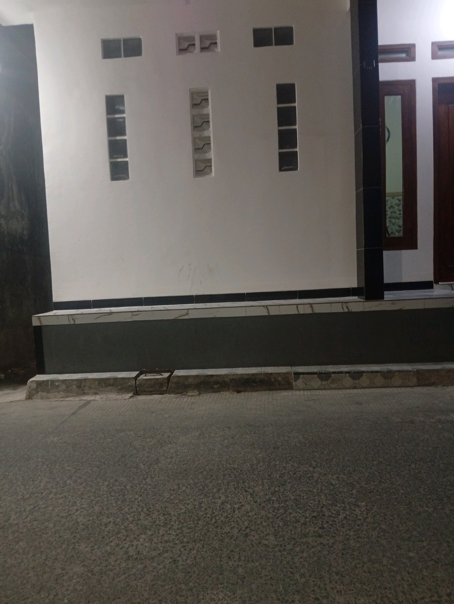 The image depicts the exterior of a building in Banowan, Indonesia. The building has a white facade with several small windows. There are at least three sets of small, rectangular windows, two with what appear to be tinted glass panes, and one set with vertical louvers. Below the windows, there is a dark gray textured base that transitions to a lighter tiled surface. In the foreground, a paved road is visible, with a concrete curb and a small section of what looks like a drainage grate. To the right of the building, a dark pillar separates two wooden doors, one of which is partially visible and appears to have a glass pane. The overall scene suggests a residential or commercial building at street level.