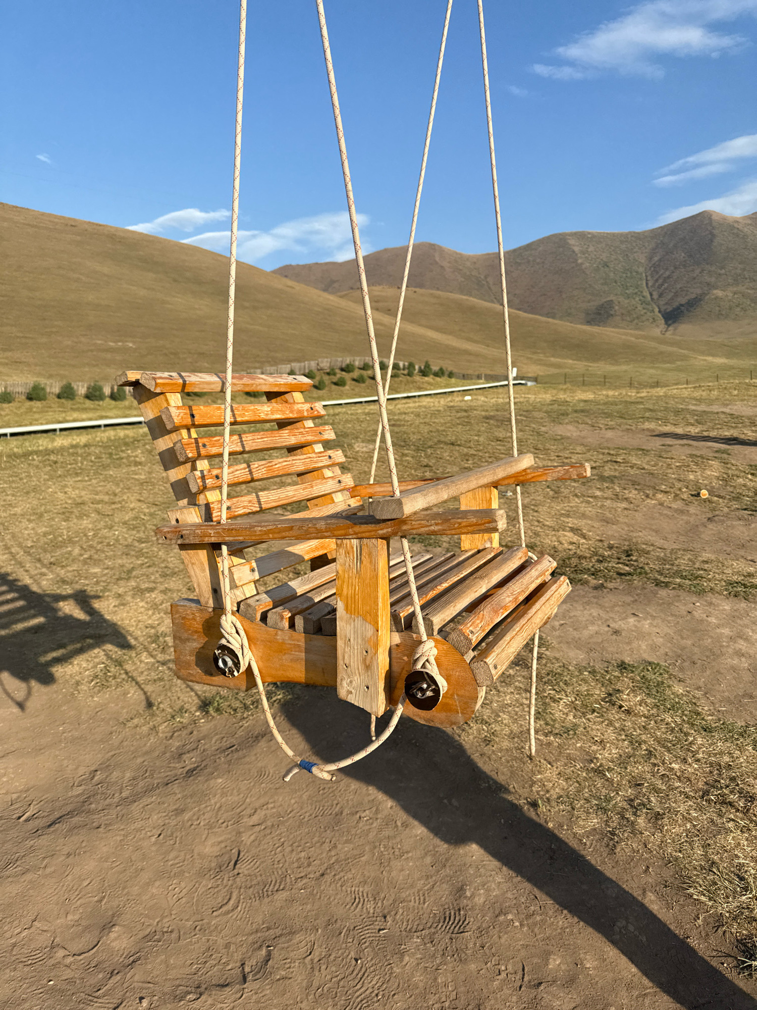 In Chunkurchak, Kyrgyzstan, a wooden swing chair is suspended by ropes against a backdrop of rolling hills and a clear blue sky. The chair, made of weathered wooden planks, is positioned in the foreground, casting a long shadow on the dusty ground. Behind the swing, the landscape transitions from dry, grassy terrain to the undulating slopes of mountains. A sparse line of trees and a white fence are visible in the middle ground, suggesting a rural or park-like setting. The lighting indicates it is late afternoon, with the sun casting a warm glow. There are no people present in the image.