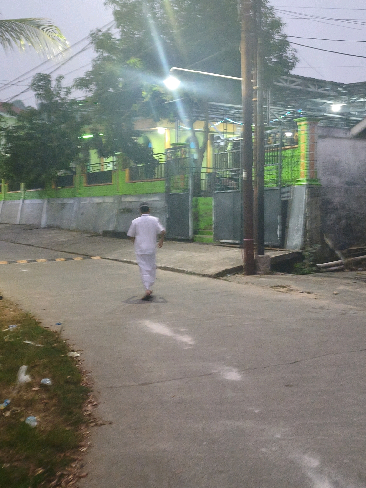 The image shows a person walking away from the camera down a street in Samarinda, Indonesia. The person is wearing a white robe and a black cap. The street is paved and has a speed bump. On the left side of the street, there is a grassy area with some trash. On the right side of the street, there is a building with a green facade and a fence. The sky is overcast. The image appears to have been taken at dusk or dawn.