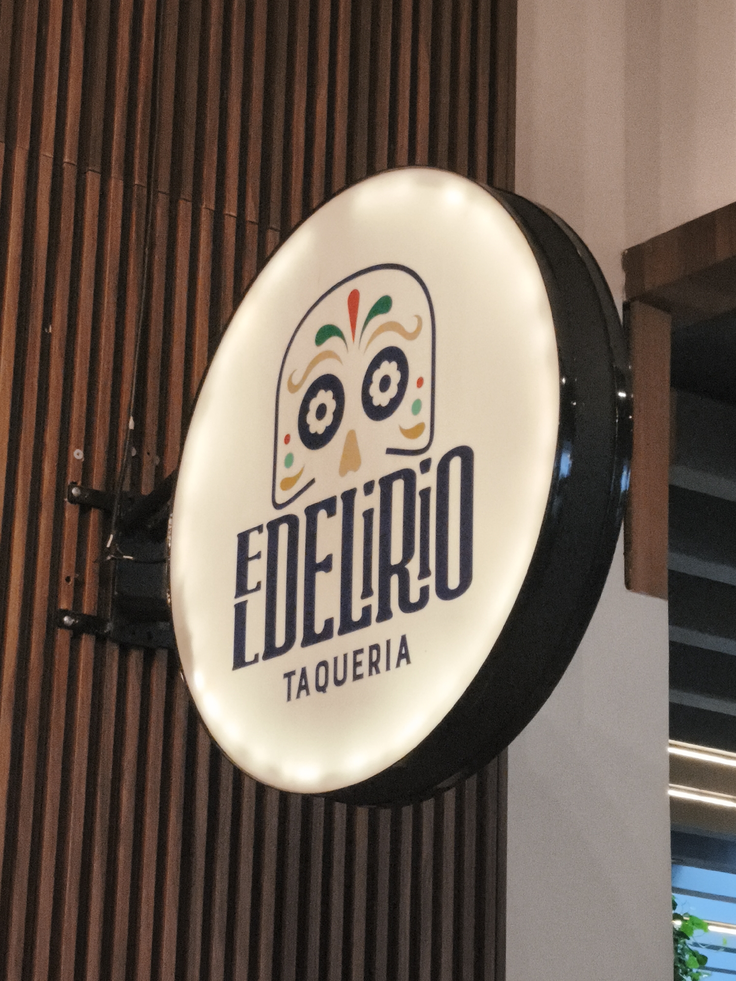 A close-up shot depicts a circular, illuminated sign mounted on a wall with vertical wood slats. The sign is white with a black rim and features a stylized skull design at the top, decorated with red, green, and yellow elements. Below the skull, the name "EDELIRIO" is printed in large, navy blue letters, followed by "TAQUERIA" in smaller, navy blue font. The sign is affixed to the wall by a black metal bracket. Parts of a doorframe and a window can be seen behind the sign. The sign's presence suggests it is the location in Guatemala City, Guatemala.