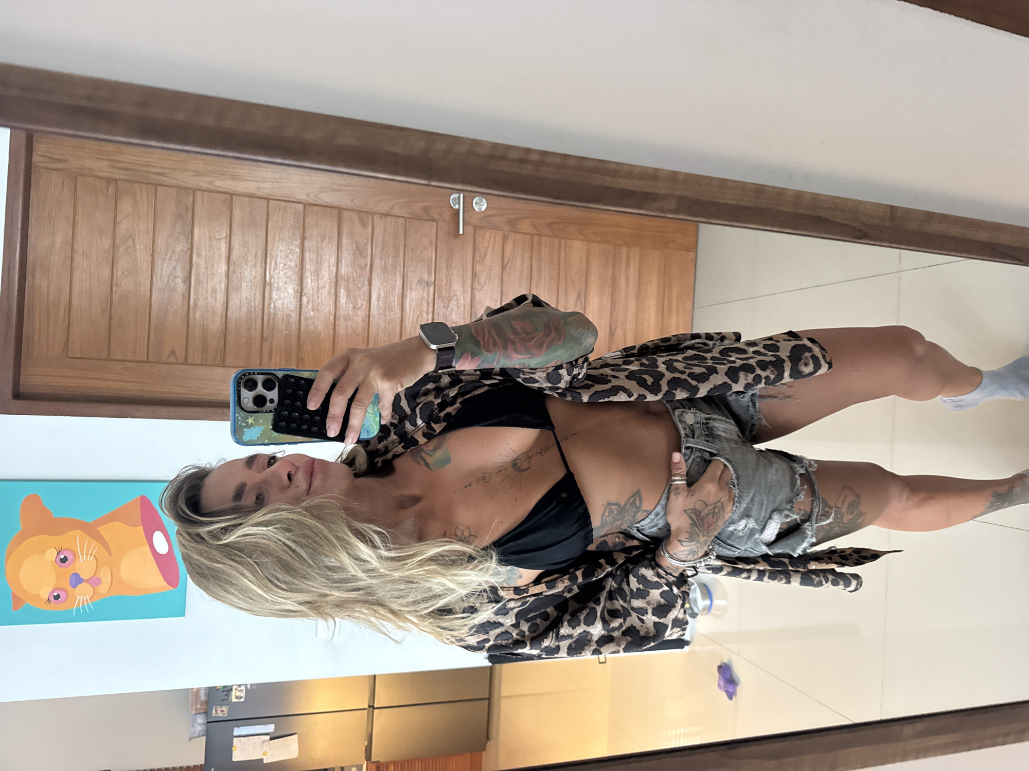 A woman is taking a mirror selfie in a brightly lit room. She has blonde, wavy hair and is wearing a black bikini top, ripped denim shorts, and an open leopard print kimono. She has multiple tattoos on her arms and torso. A smartwatch is visible on her left wrist. In the reflection, a refrigerator and a colorful painting of a cat head are visible in the background. A wooden door is also reflected in the mirror. The floor is tiled. The location is identified as a residential area in Thep Krasatti, Thailand.
